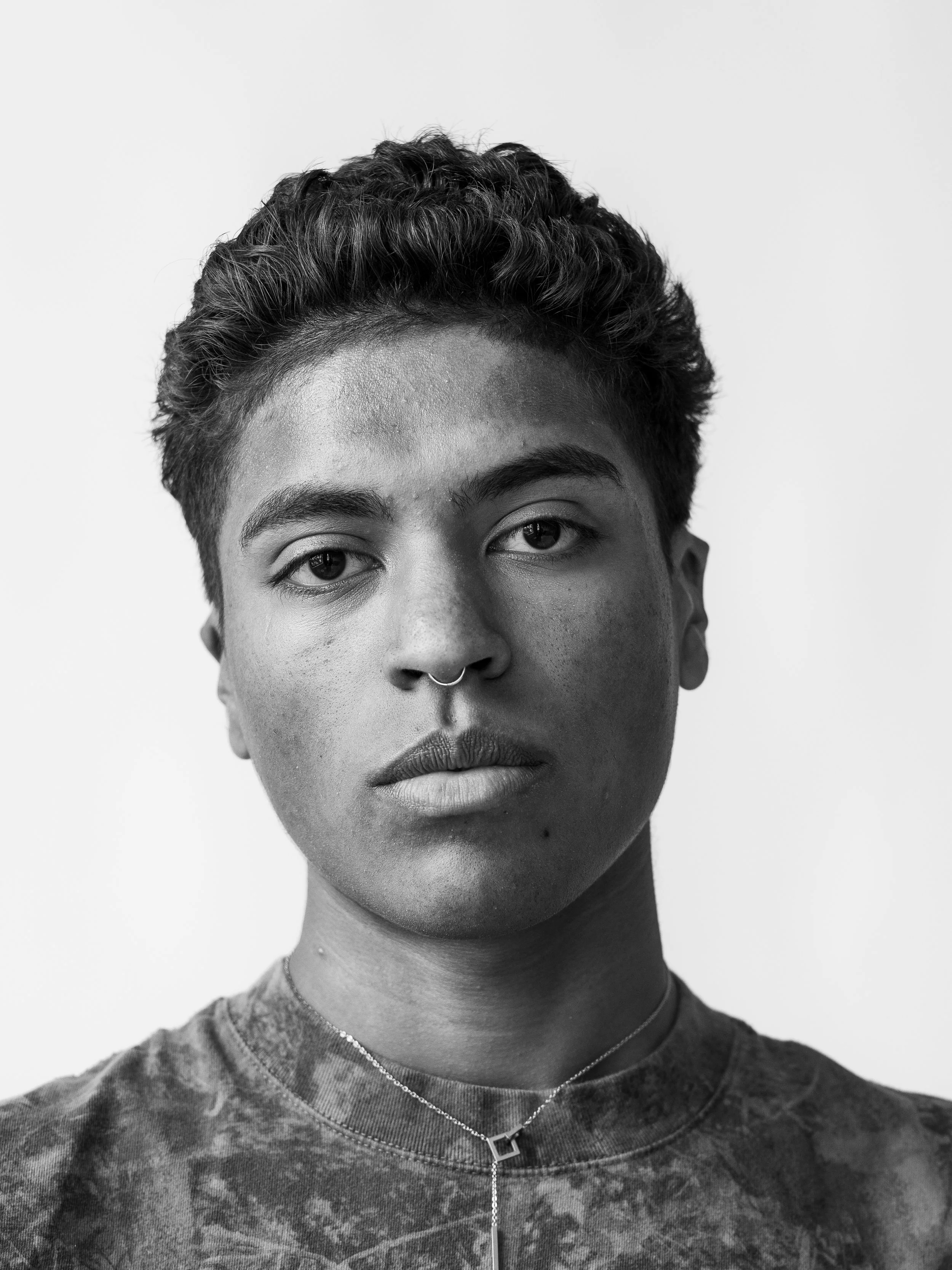 Black and white portrait of a person with short, curly hair, wearing a patterned shirt and a necklace with a square pendant and a chain, facing the camera with a neutral expression.