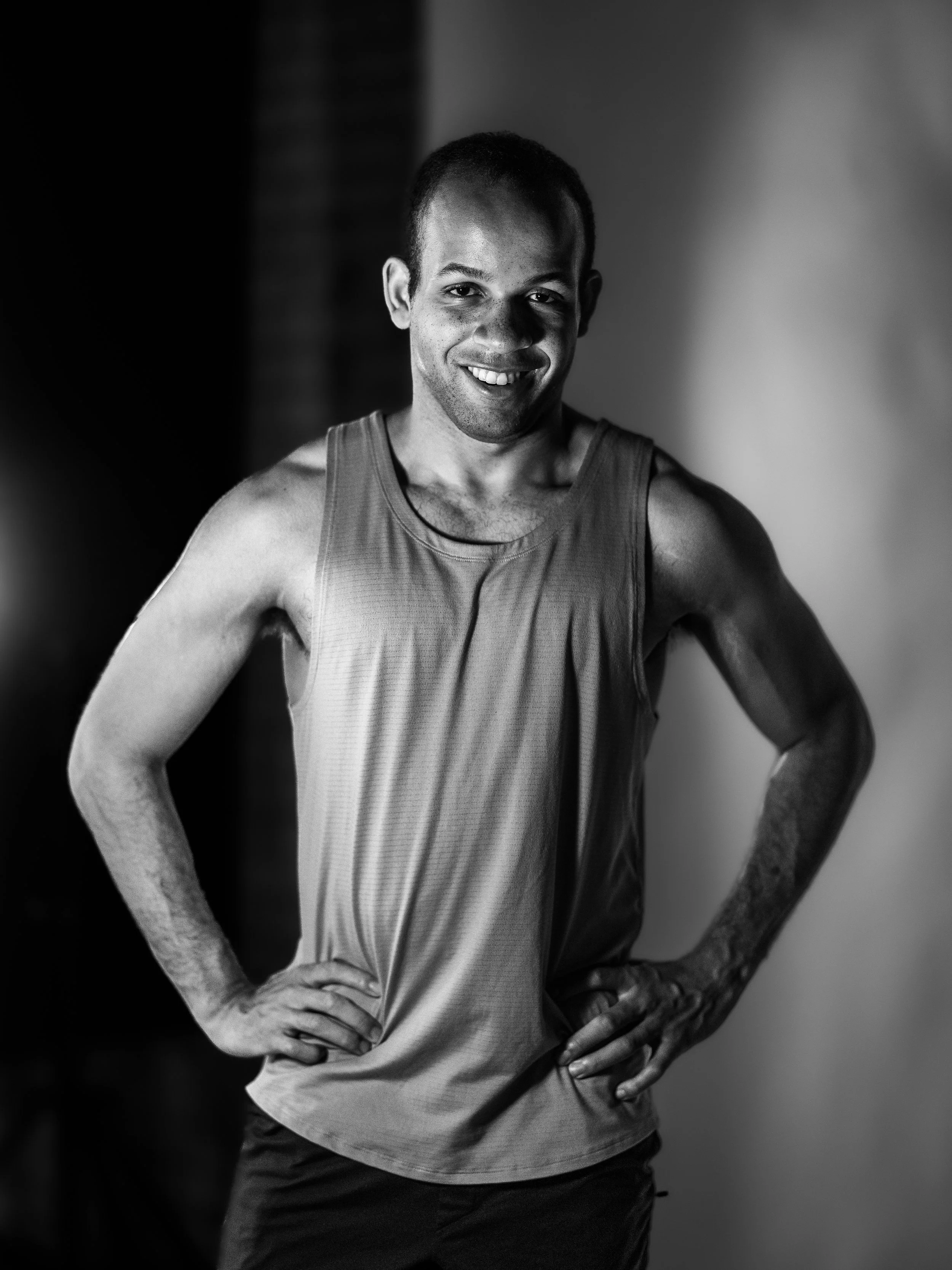 A man smiling with hands on hips, wearing a sleeveless athletic shirt, in black and white photography.