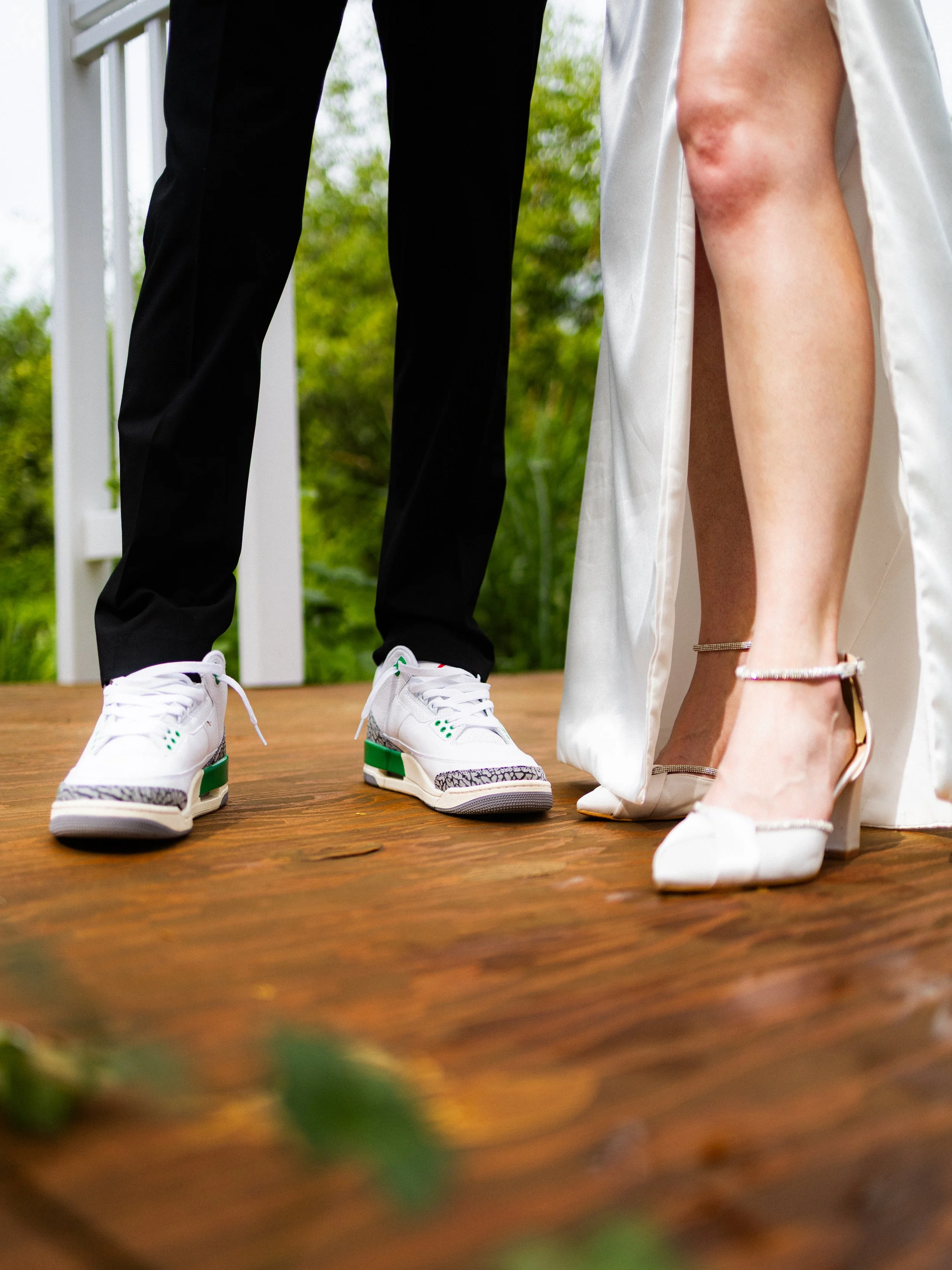 Close-up of a person's legs, one in a wedding dress showing a high-heeled shoe, the other in black pants with sneakers, standing on a wooden deck.