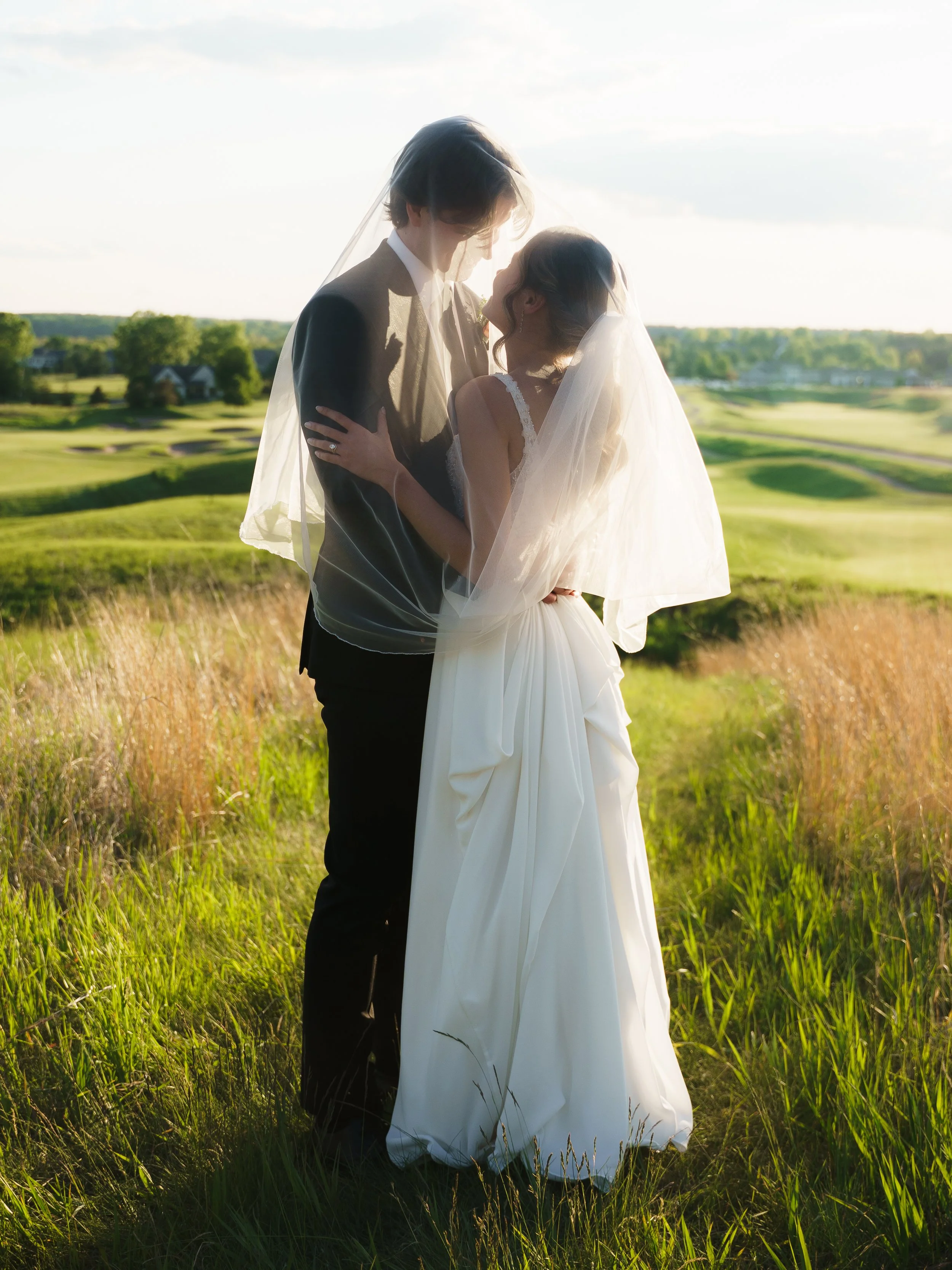 A bride and groom standing close together outdoors, holding each other, with a scenic green landscape and golf course in the background during sunset.