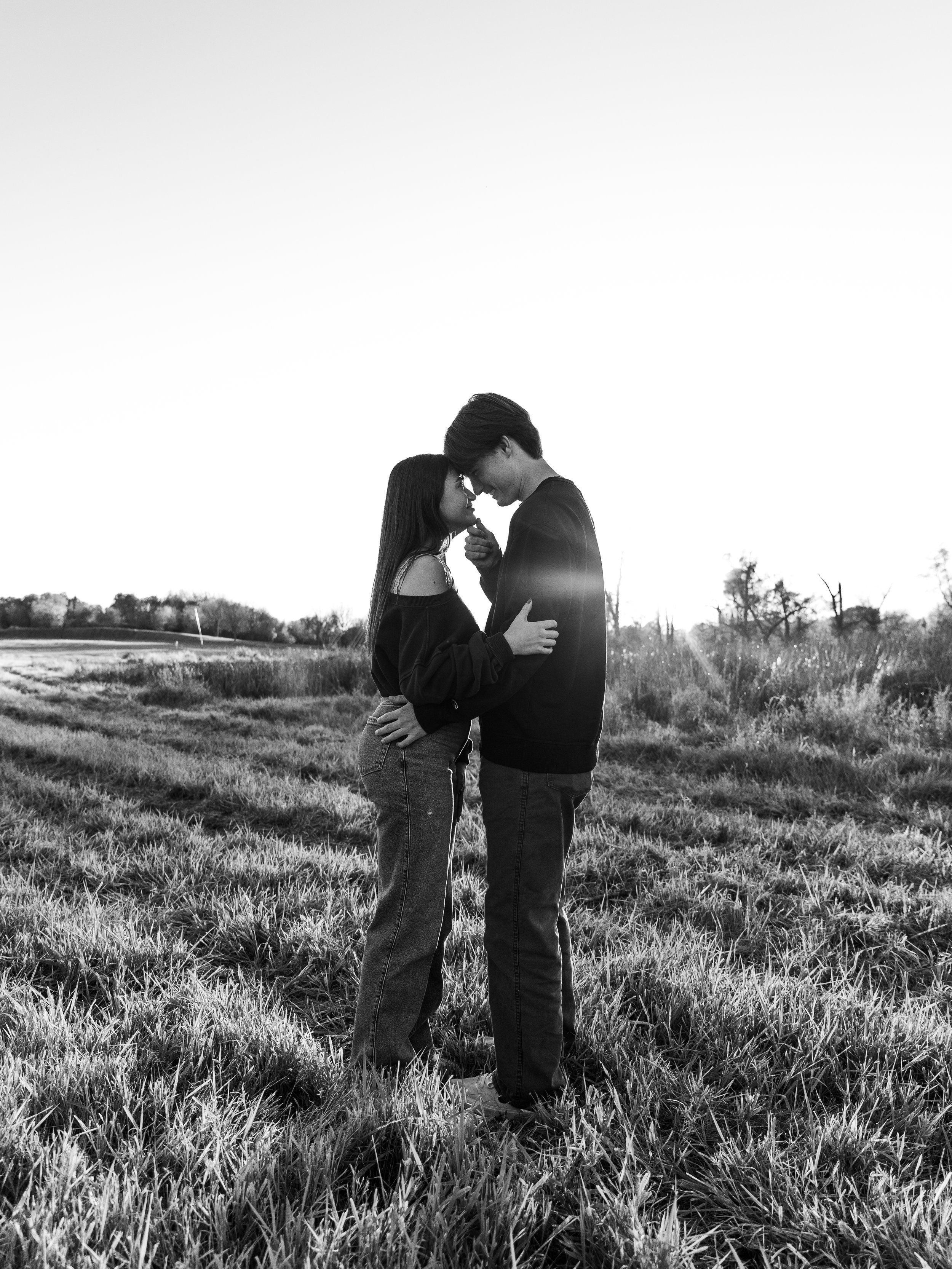 A black-and-white photo of a couple standing close together in a grassy field at sunset, touching foreheads and smiling.
