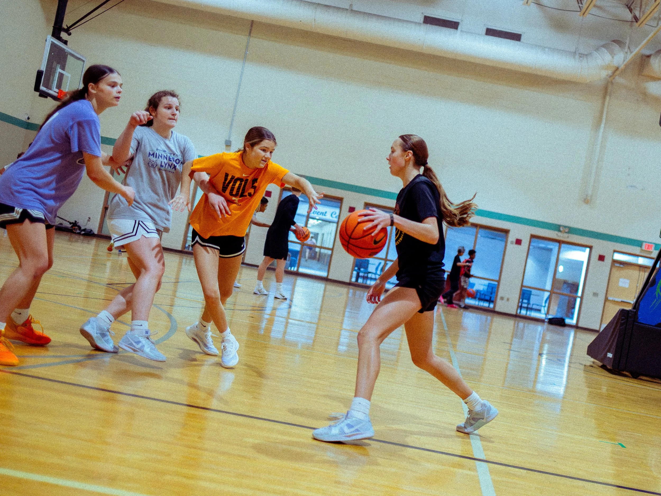 Four young women playing basketball in an indoor gym, with one girl dribbling the ball while the others defend or prepare to receive a pass.