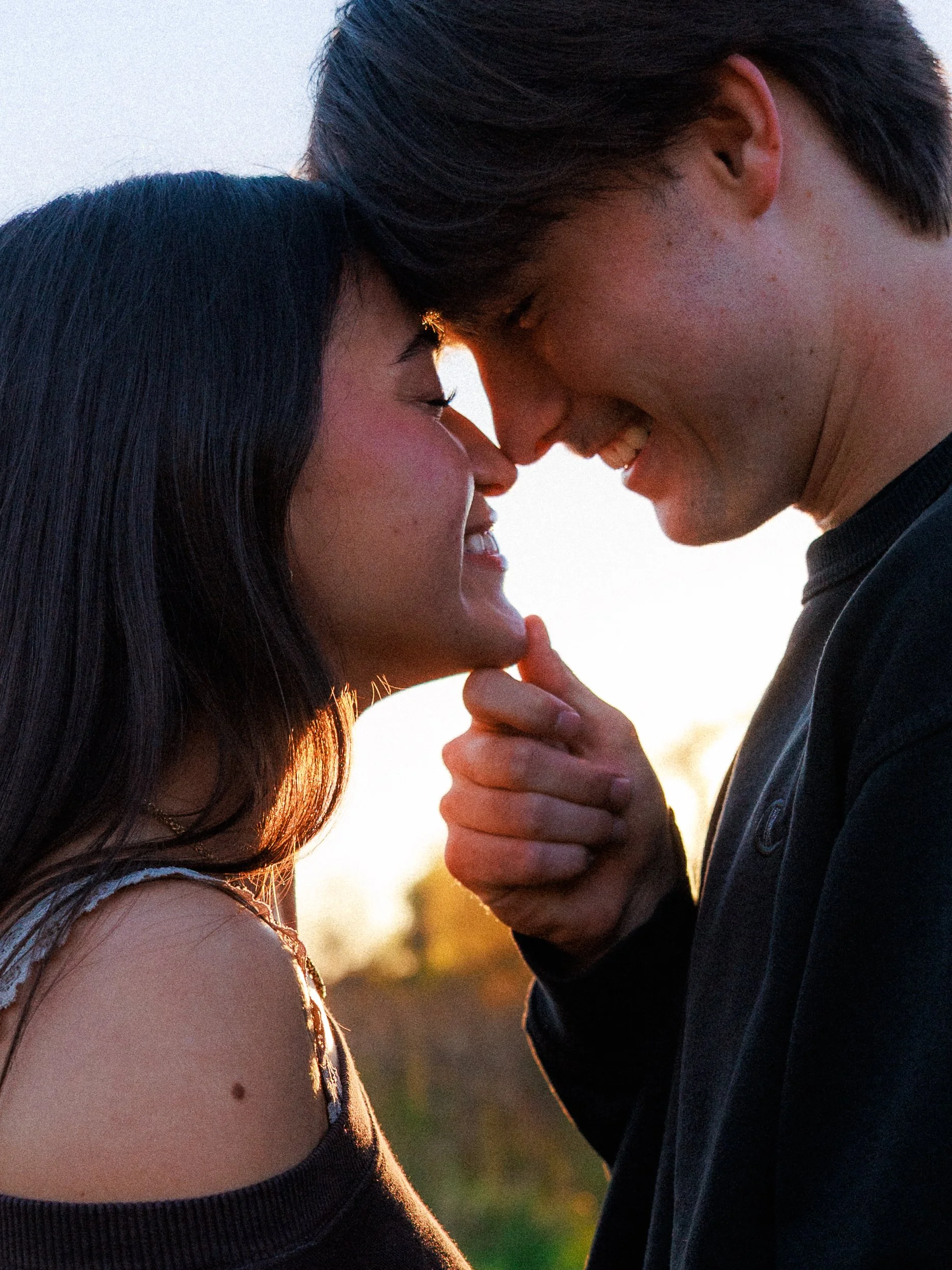 A couple faces close together outside during sunset, smiling and touching foreheads, with the man gently touching the woman's chin.