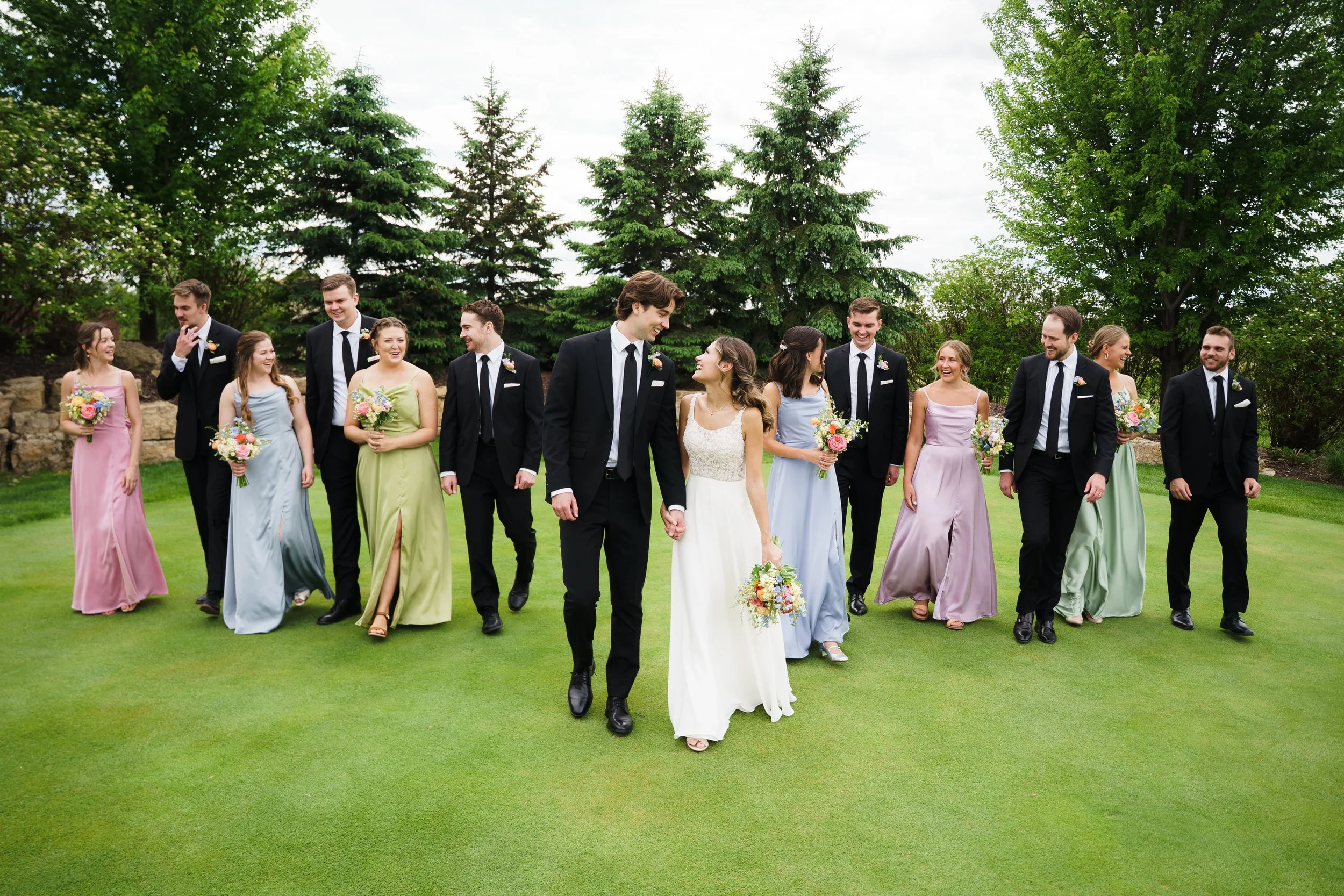A wedding party walking on a lush green lawn with trees in the background, featuring the bride and groom in the center holding hands, surrounded by bridesmaids in pastel dresses and groomsmen in black suits, all smiling and enjoying the moment.