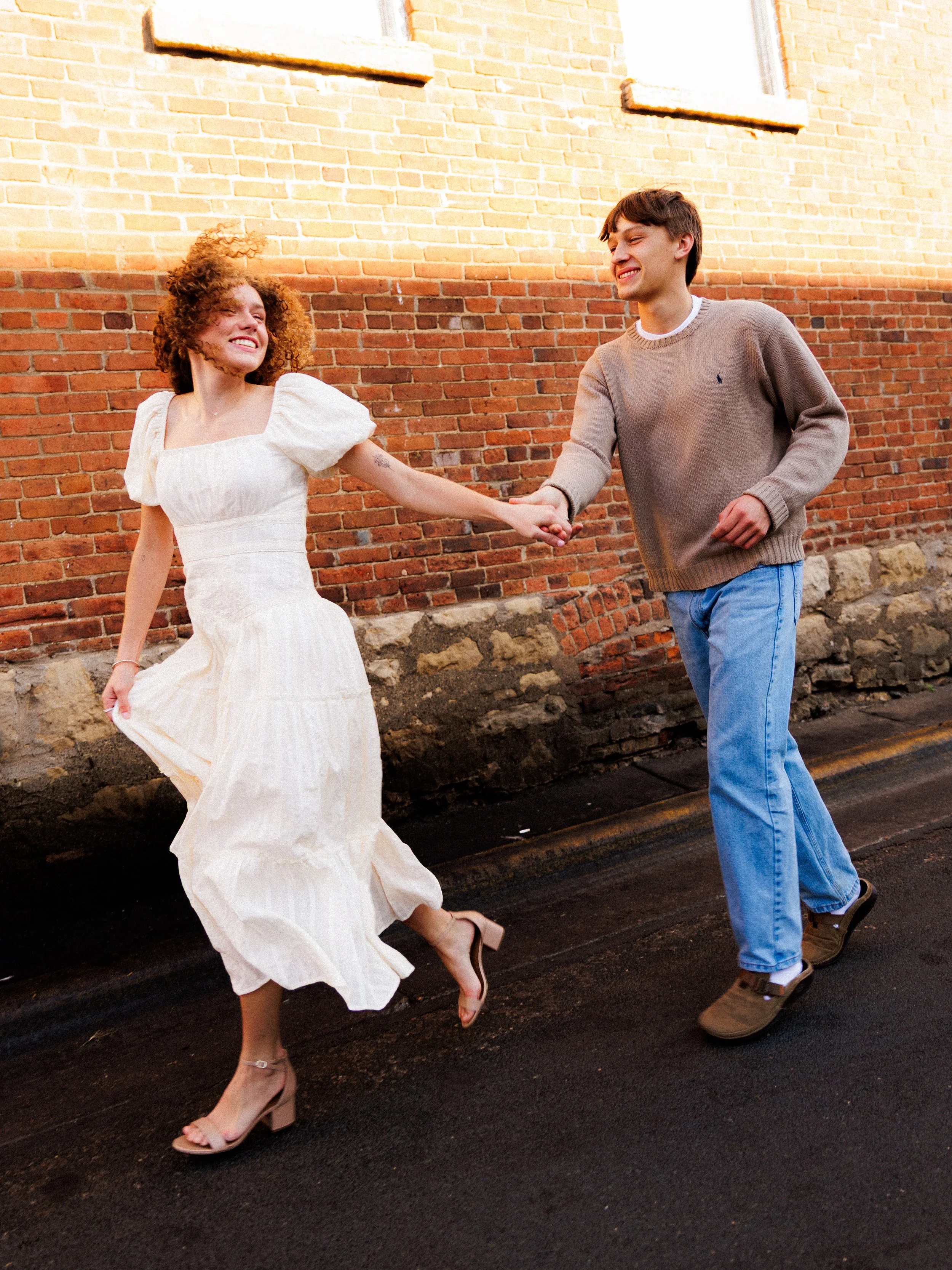 A young couple holding hands and smiling while dancing on the sidewalk in front of a brick building.