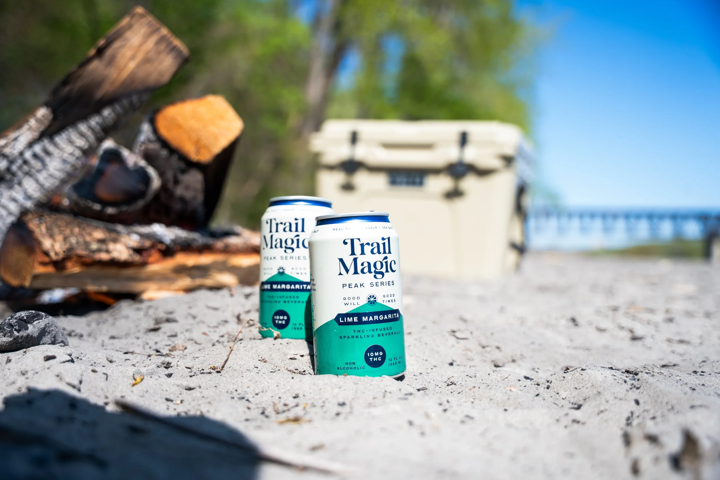 Two cans of Trail Magic Lime Margarita sparkling beverage with THC infusion on sandy ground near a campfire with logs and a cooler in the background.