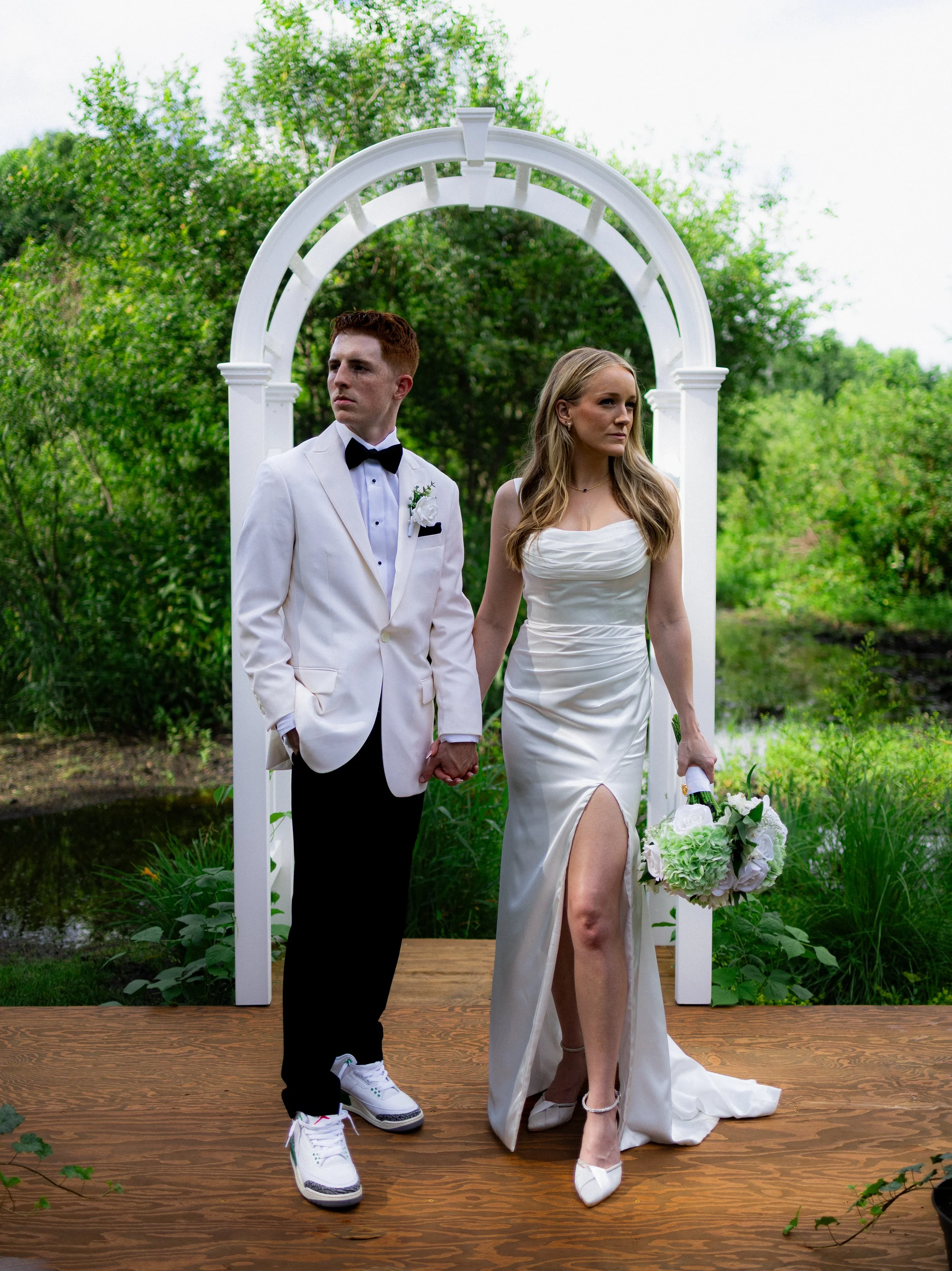 A bride and groom standing hand in hand on a wooden platform outdoors, with a white arch and lush green trees in the background. The bride is in a white dress with a thigh-high slit, holding a white bouquet, and the groom is in a white tuxedo jacket 