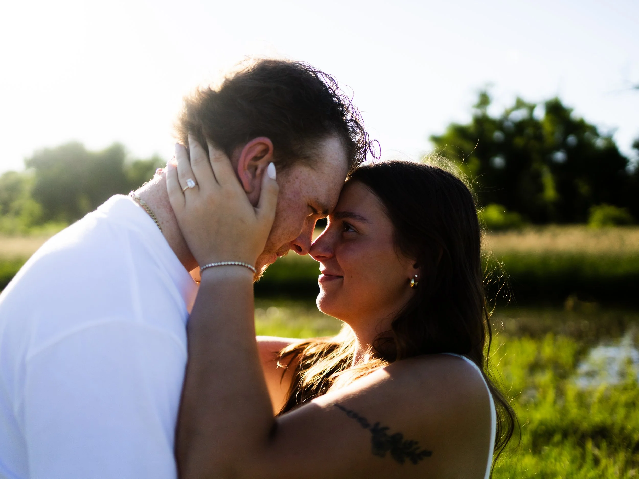 A couple faces close together outdoors with a river and trees in the background.