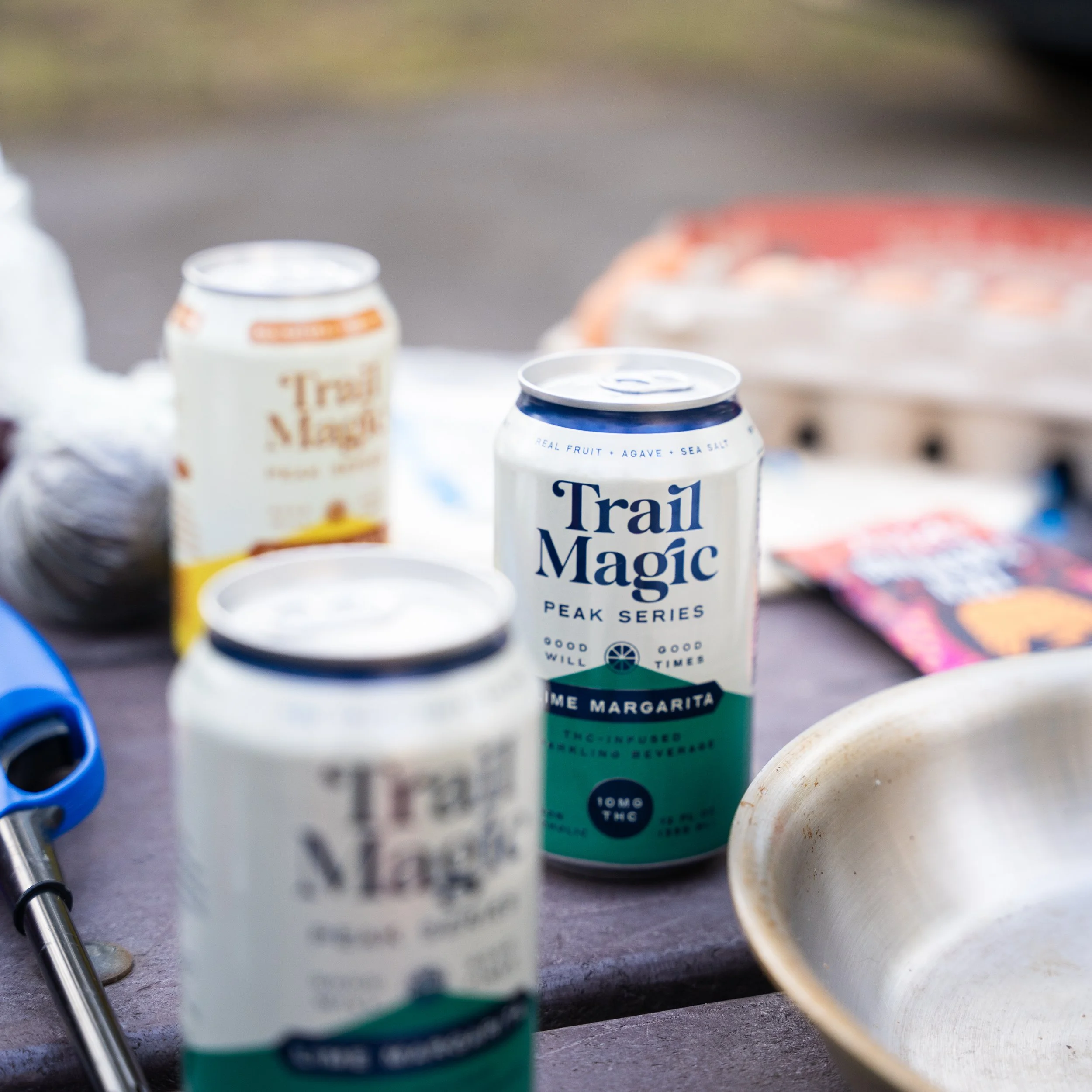 Three cans of Trail Magic Peak Series seltzer water on a table, with a metal bowl and other items around, outdoors.