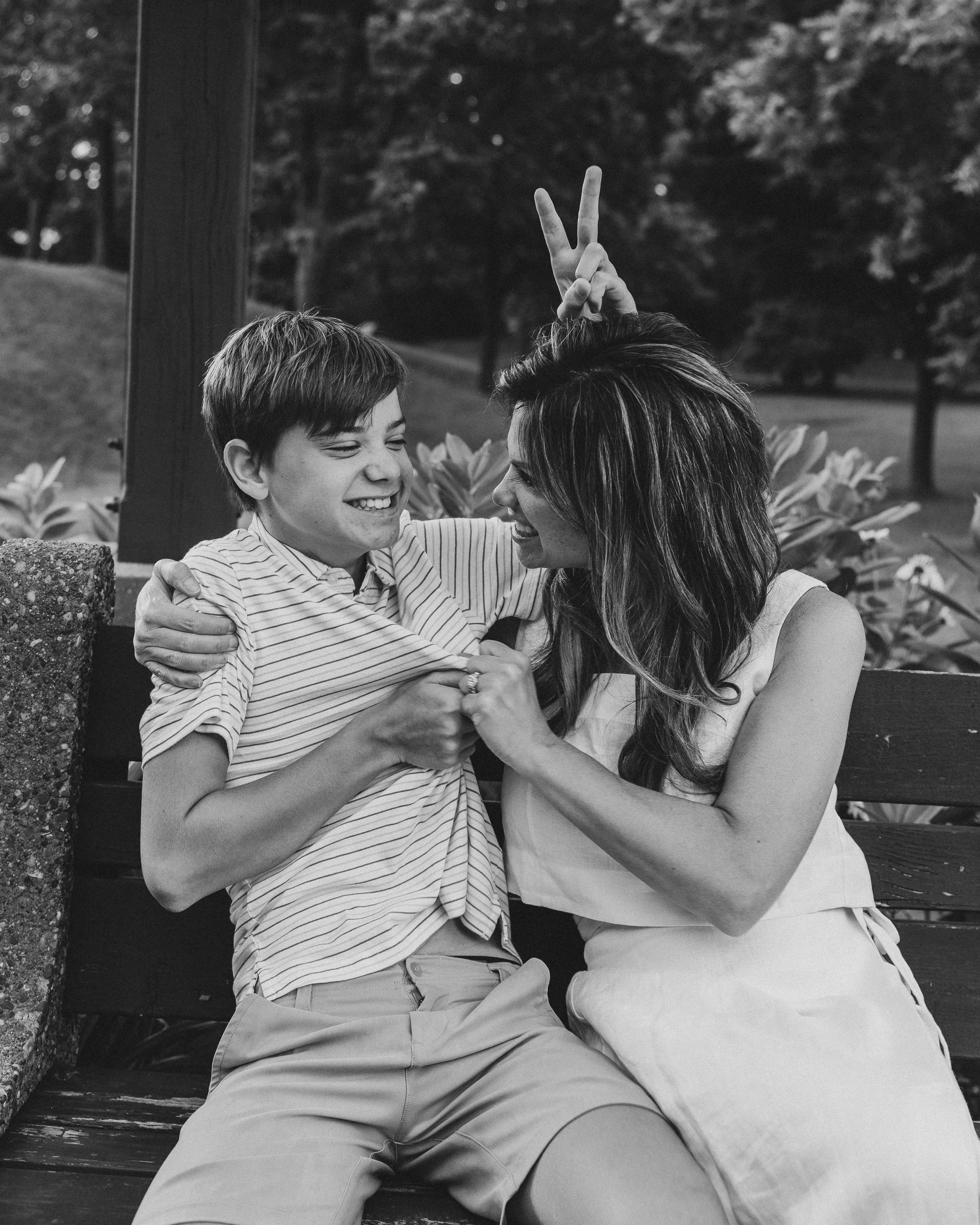 A woman and a boy smiling on a park bench. The woman playfully makes bunny ears behind the boy's head. They are holding each other and appear to be enjoying a happy moment together.