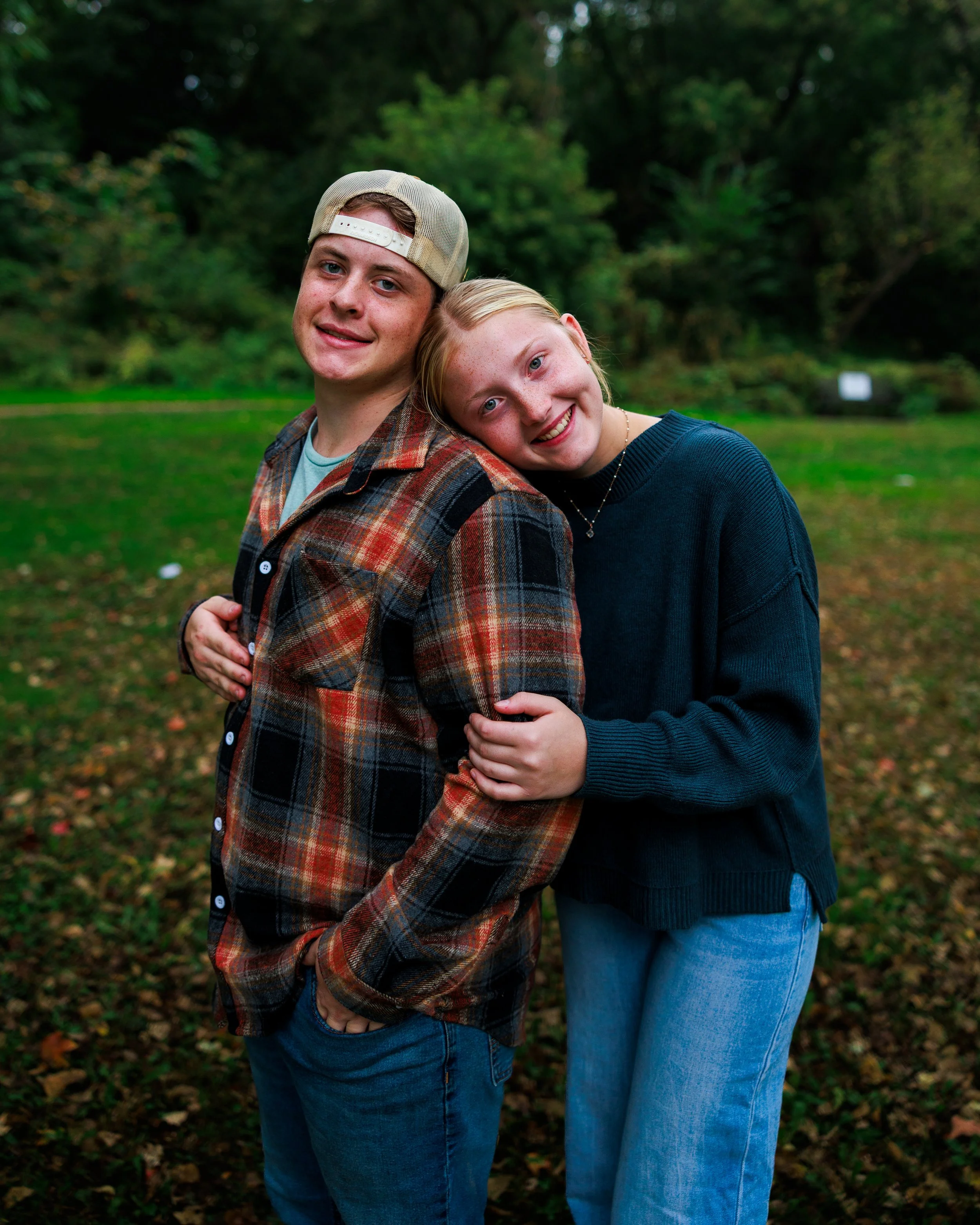 A young man and woman are standing outdoors, smiling, with the woman hugging the man's shoulder.
