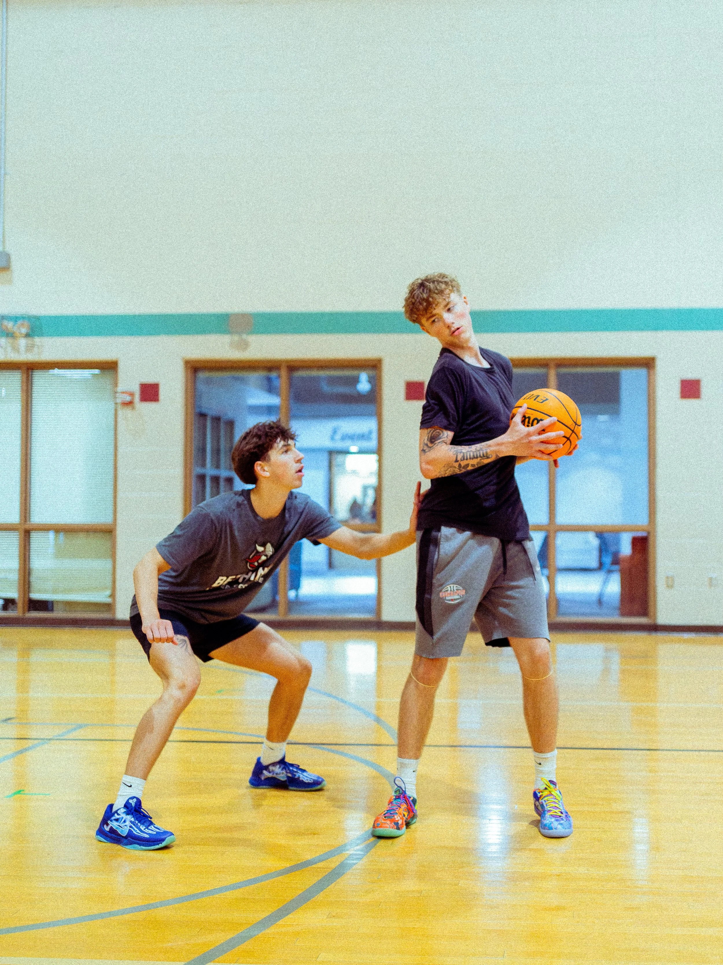 Two young men playing basketball in an indoor gym; one standing with a basketball, the other crouching defending.