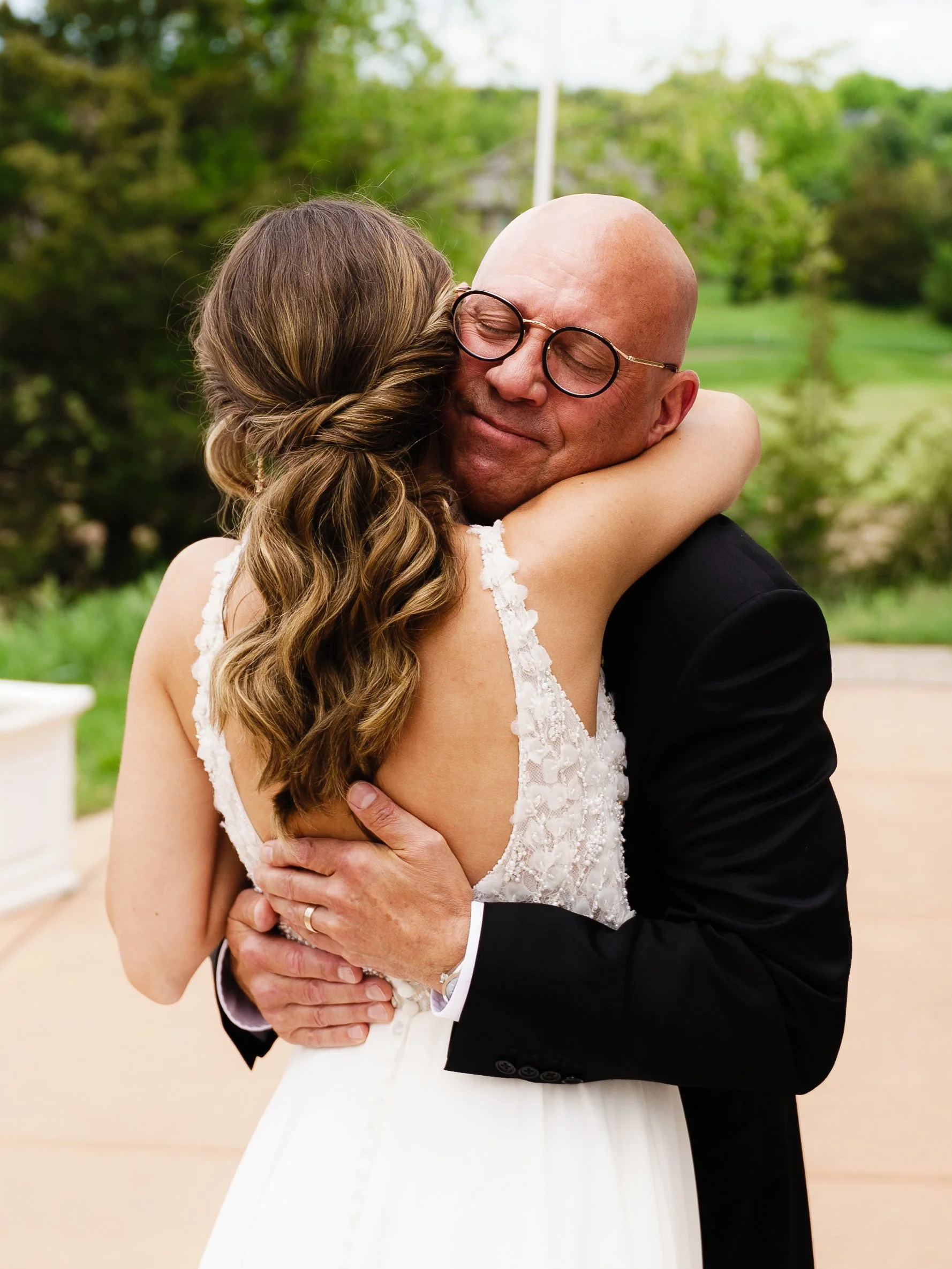 A woman in a wedding dress hugging a bald man in glasses and a black suit outdoors with trees and greenery in the background.
