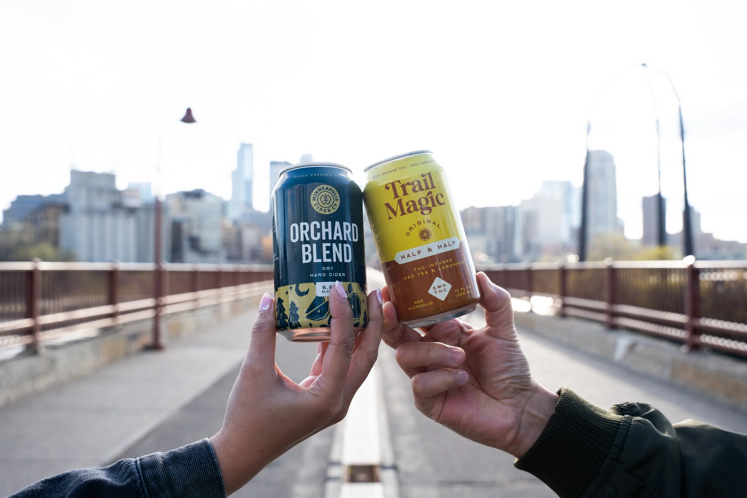 Two people holding canned beverages, one Orchard Blend and one Trail Magic, against an urban cityscape background with a bridge and tall buildings.