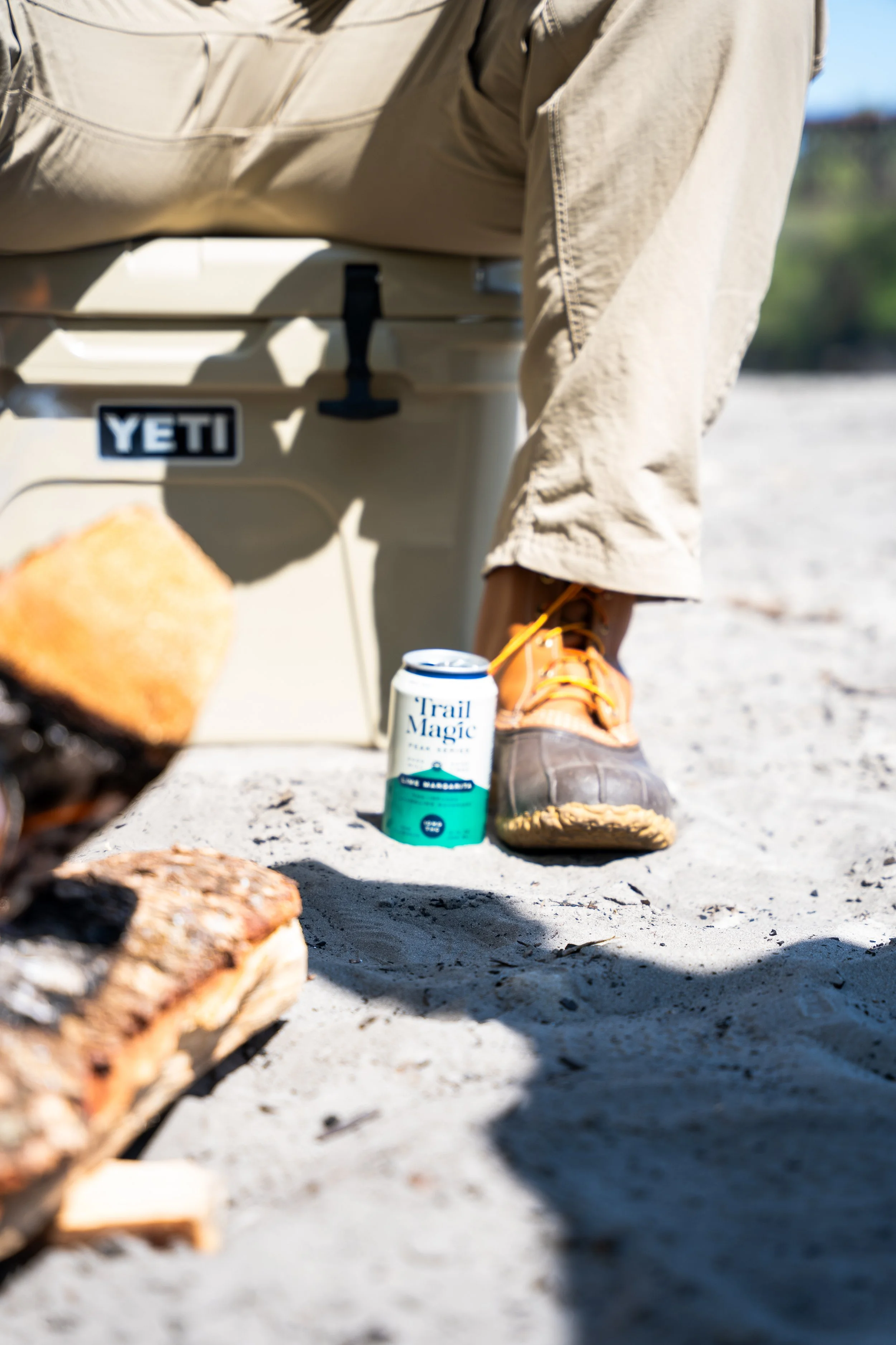 A can of Trail Magic beer on sandy ground near a person's leg and a YETI cooler in the background.