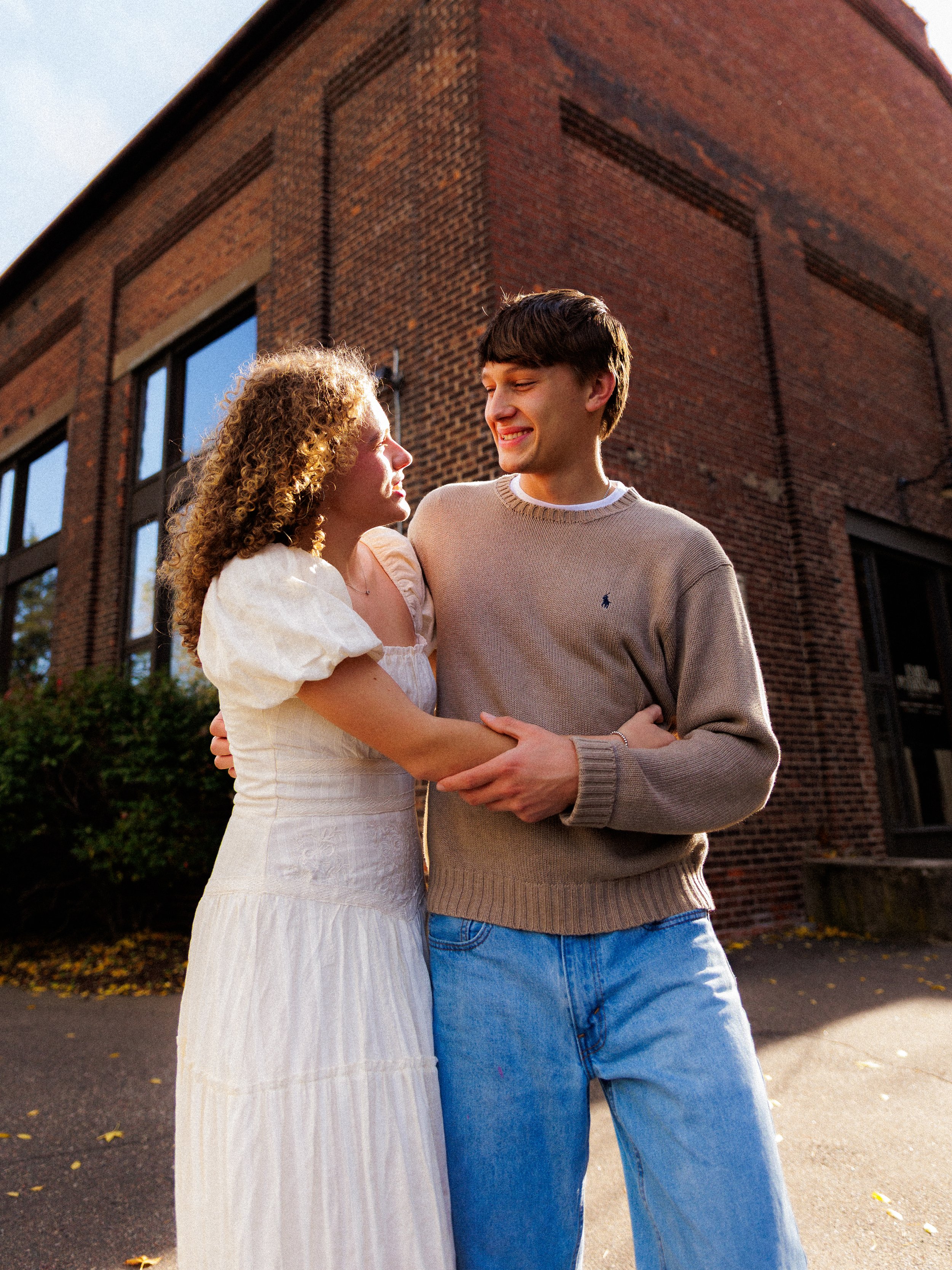 A young man and woman are smiling and holding each other outside in front of a brick building, enjoying a sunny day.