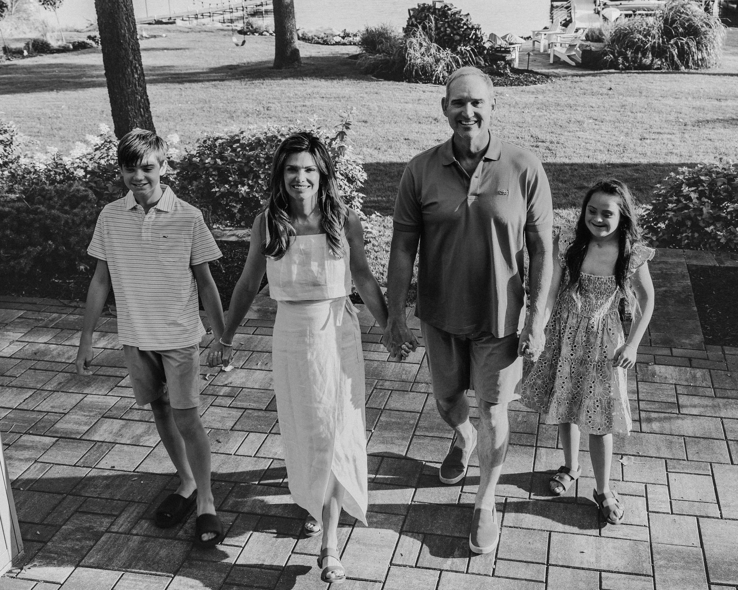 A family of five walking outdoors on a paved path, holding hands and smiling. The background features trees, bushes, and a grassy yard.