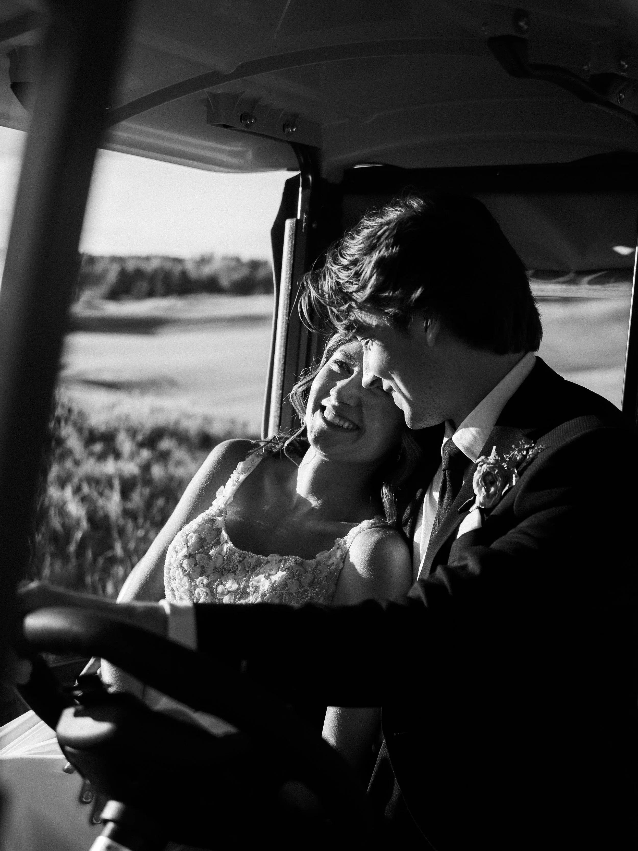 A smiling woman in a wedding dress and a man in a tuxedo sit closely together in a golf cart, sharing an intimate moment outdoors.