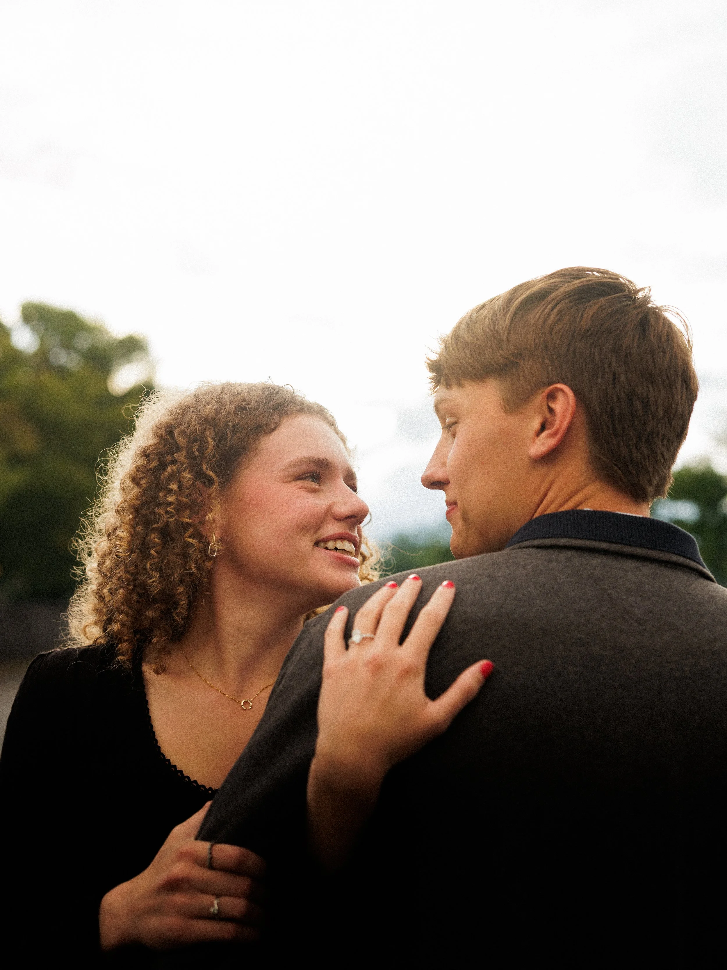 A woman with curly hair and a man with light brown hair share a close moment outdoors, smiling at each other with a blurred natural background.