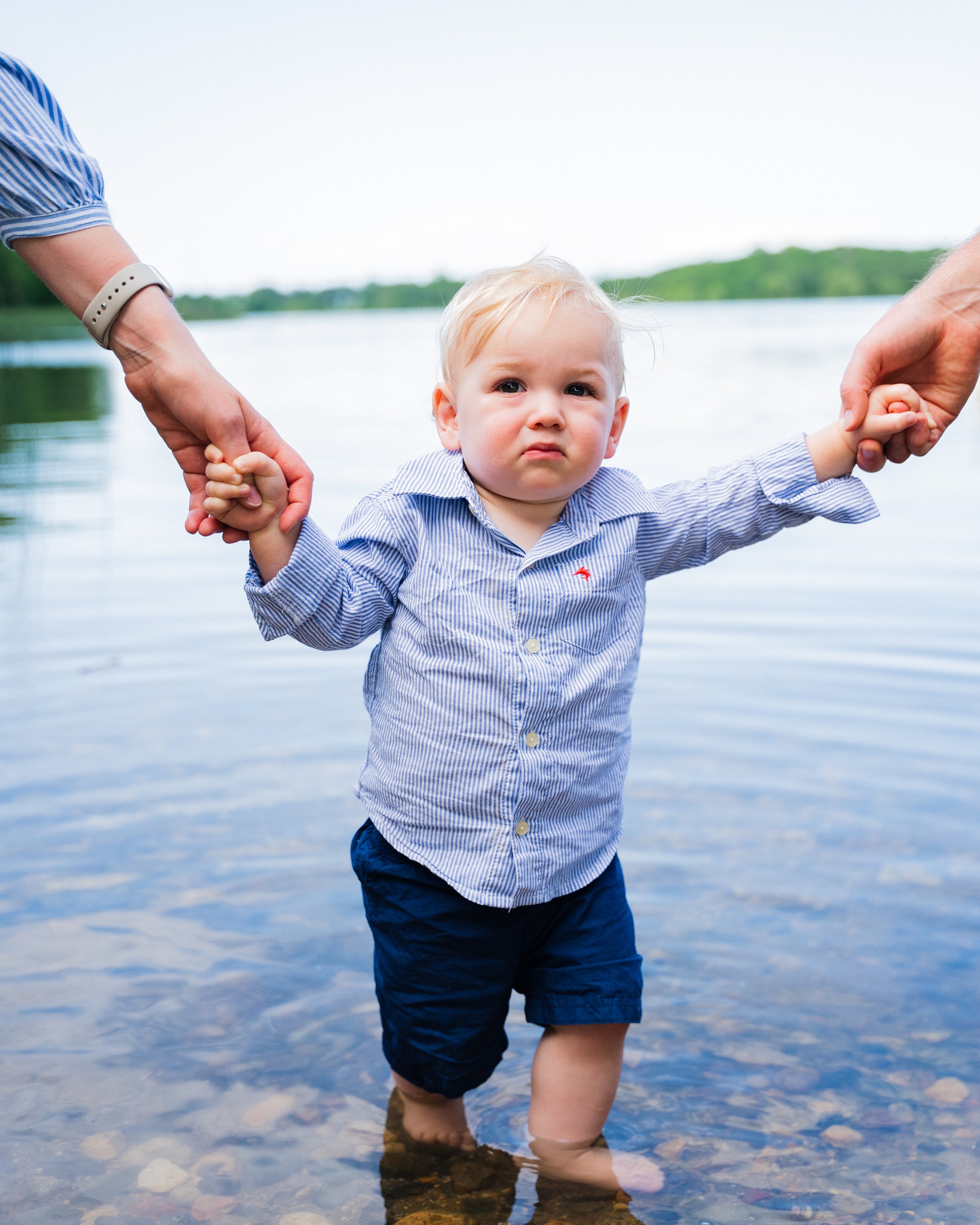 Young child standing in shallow water at a lake, holding hands with two adults, looking unhappy or upset.