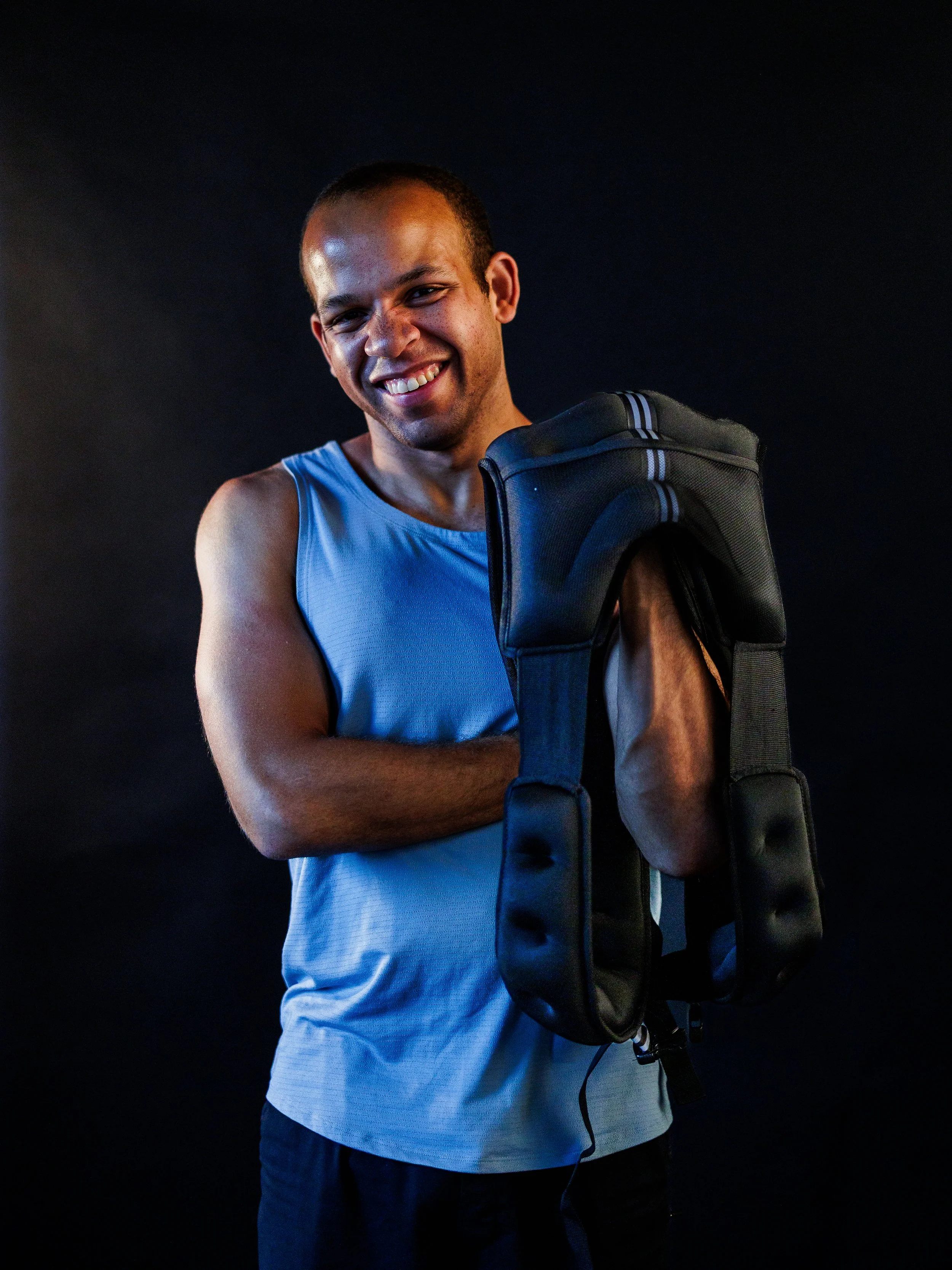 A smiling man in a blue sleeveless athletic shirt holding a black tennis bag over his shoulder, standing against a dark background.