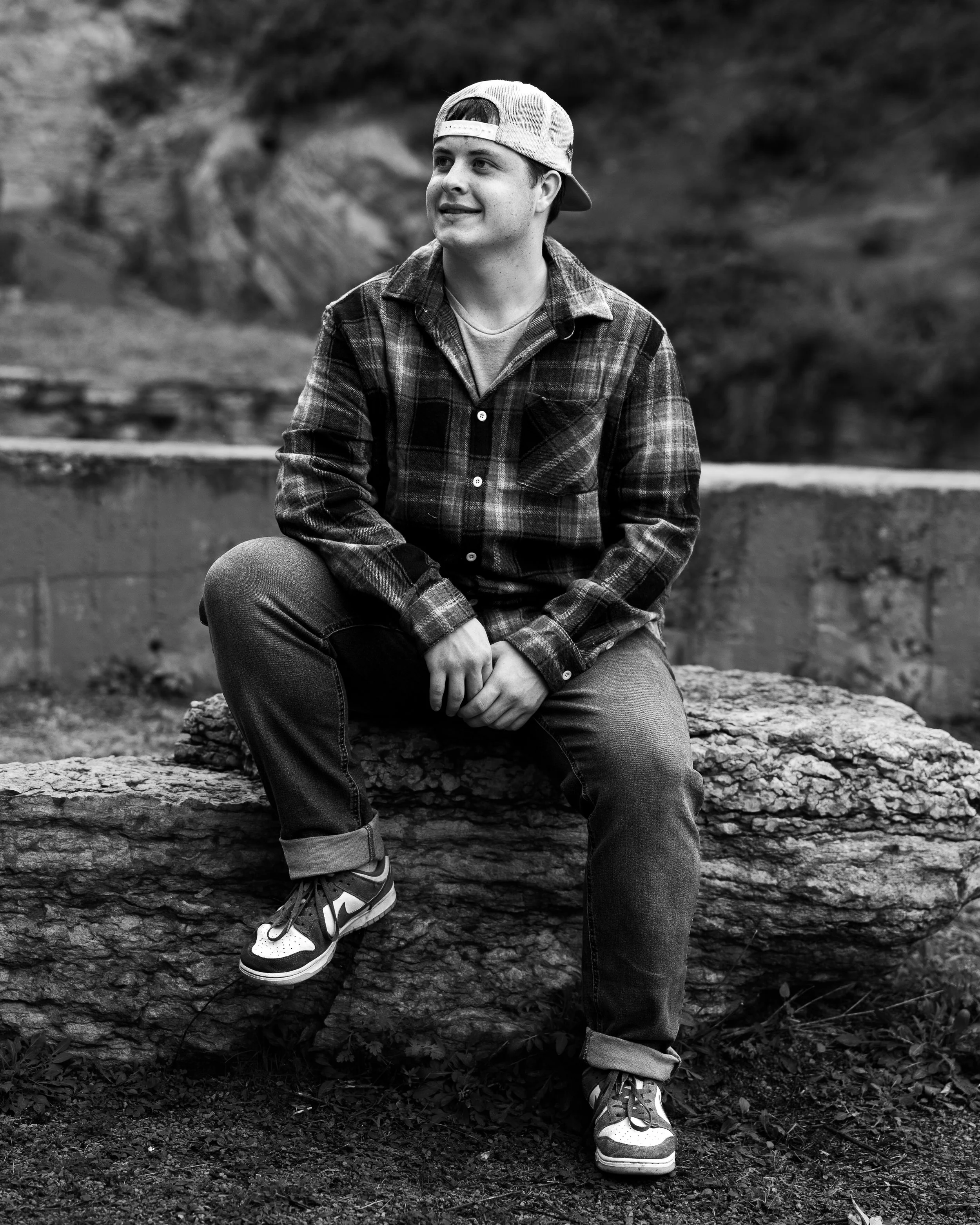 A young man in a backward baseball cap, plaid shirt, jeans, and sneakers sitting on a large rock outdoors with a blurred natural background.