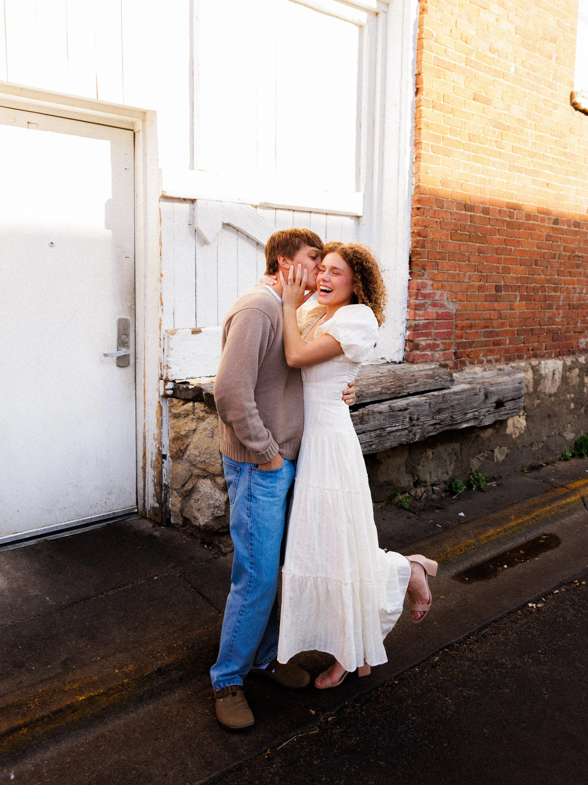 Two young people, a man and a woman, sharing a joyful moment on a sidewalk outside a brick building, embracing and laughing.