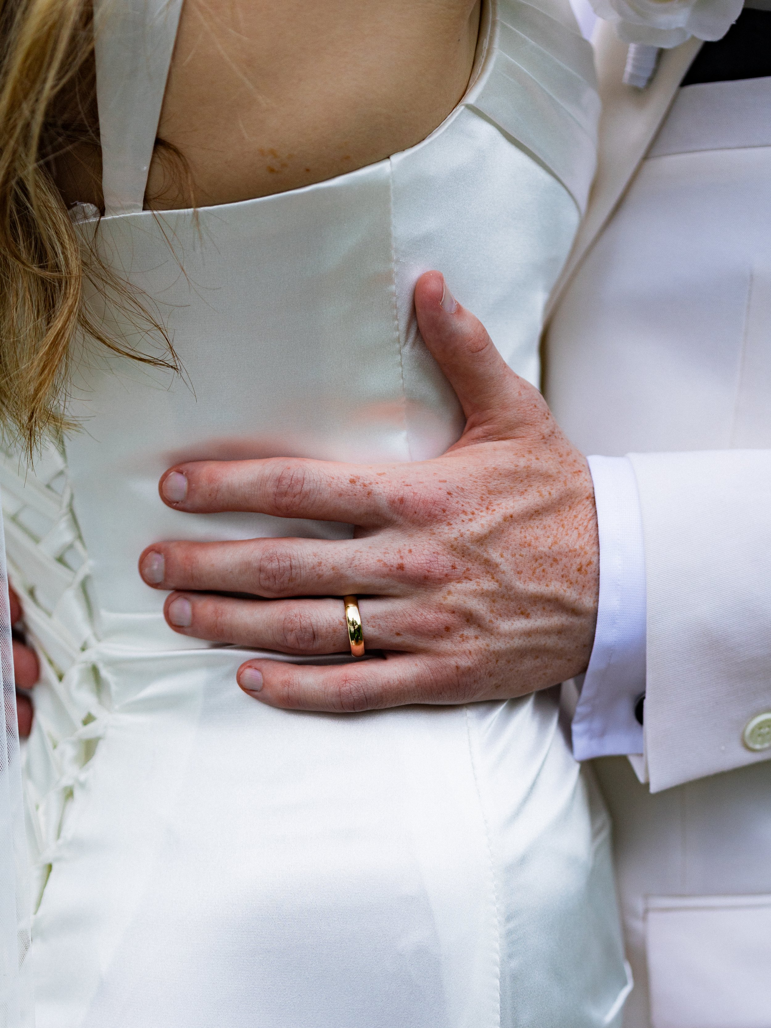 Close-up of a person's hand with a wedding band resting on the waist of someone wearing a white satin wedding dress, with a man's arm in a white suit sleeve visible.