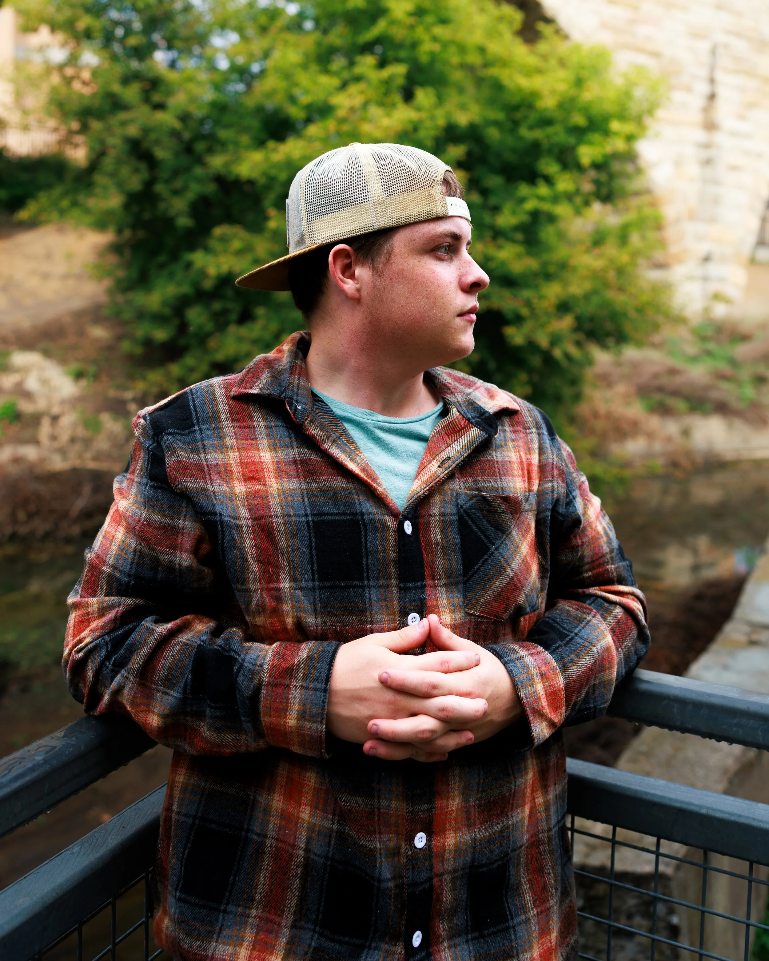 A young man with fair skin and short brown hair wearing a light-colored trucker cap, a teal T-shirt, and a red plaid flannel shirt, standing on a bridge with a black railing next to a body of water, looking to the right with a thoughtful expression, 