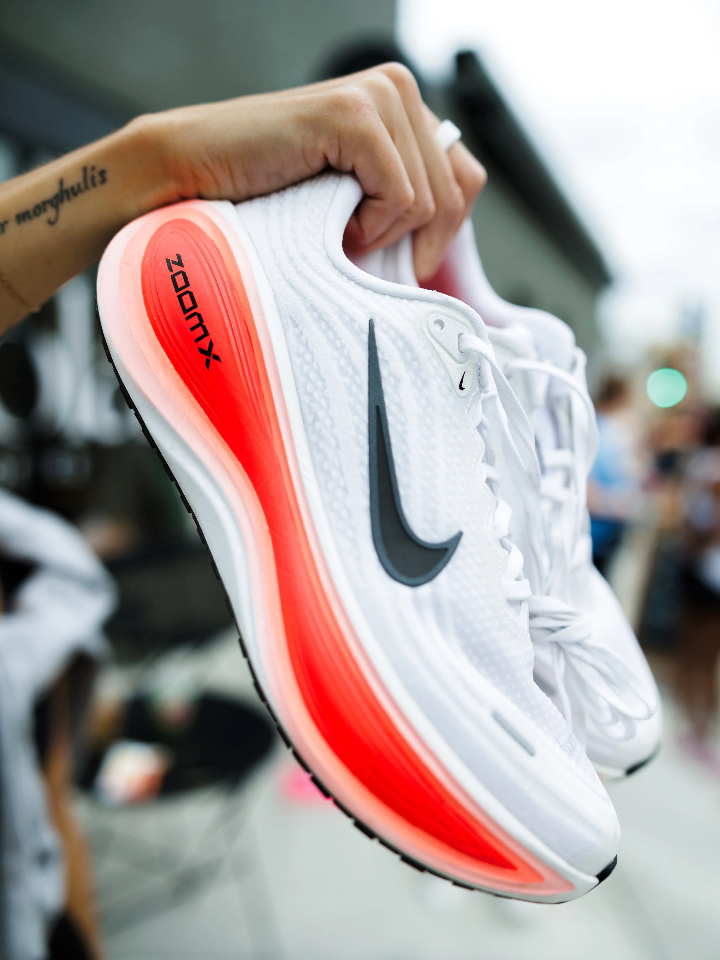 Close-up of a white Nike running shoe with black swoosh logo and a red and pink gradient Air zoom sole, being held by a person with tattooed arm.