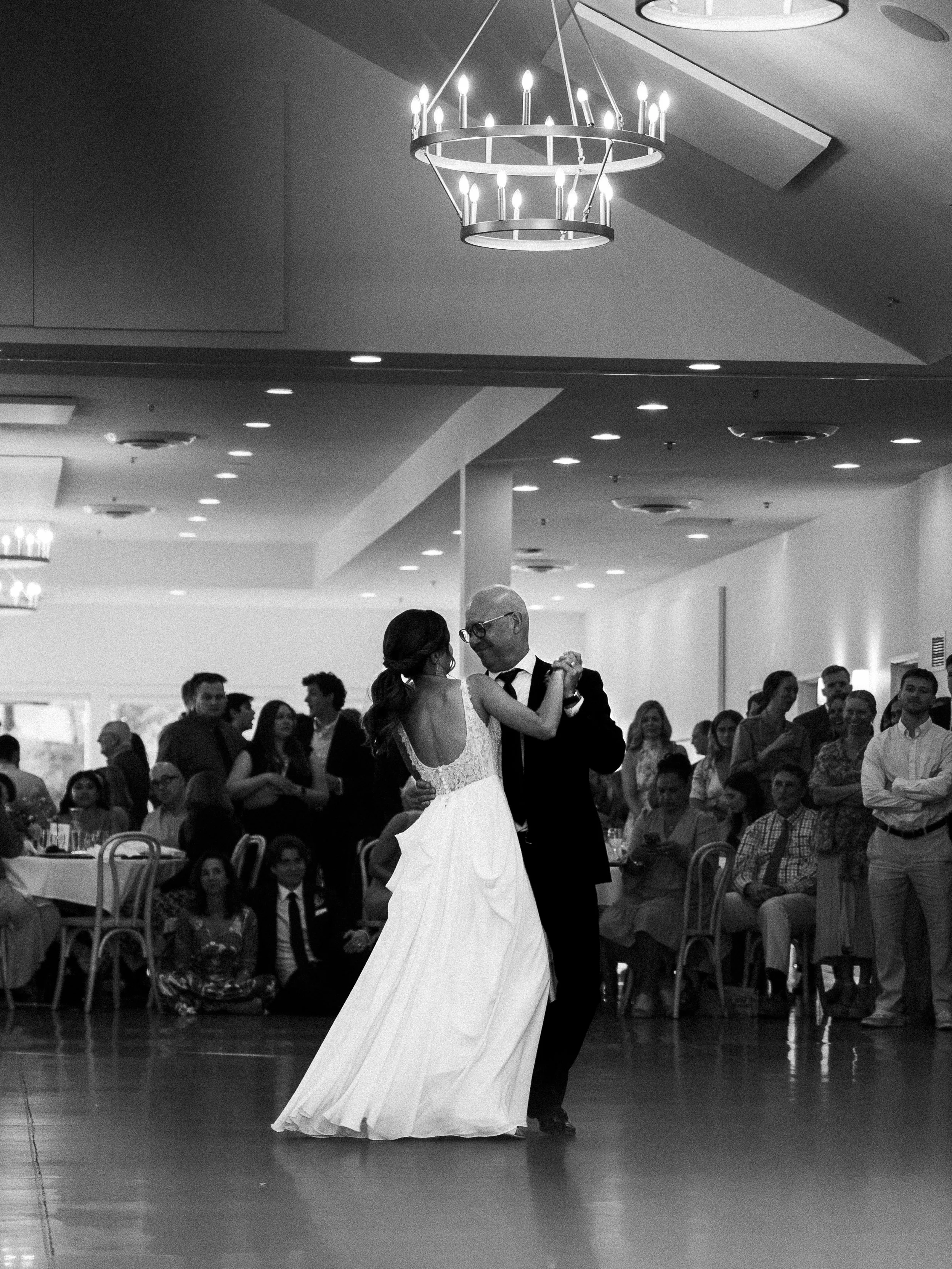Black and white photo of a couple dancing at a wedding reception, with guests watching in the background.