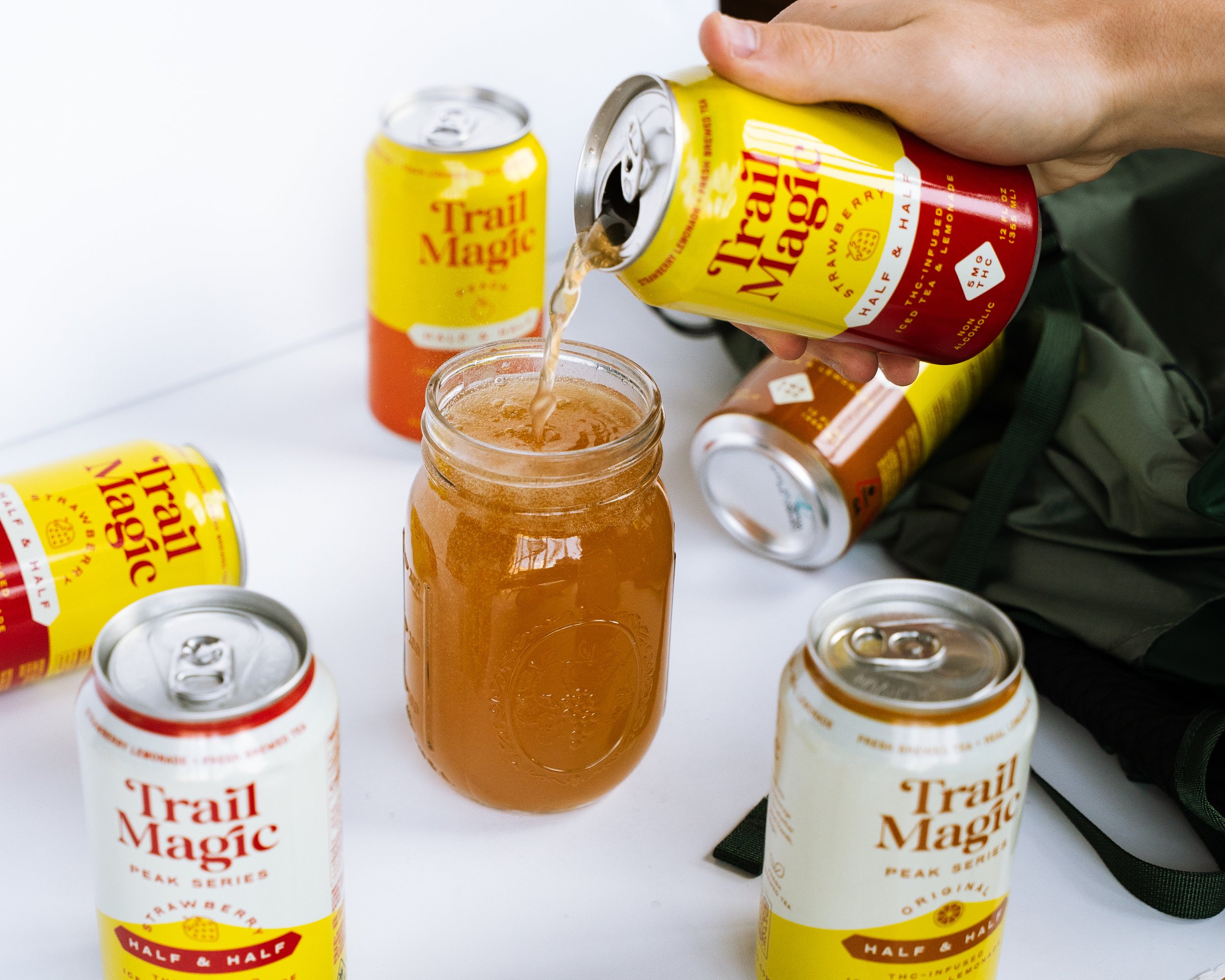 Person pouring Trail Magic sparkling beverage from can into glass jar, with multiple cans of Trail Magic drinks on a white surface, and a dark green backpack nearby.