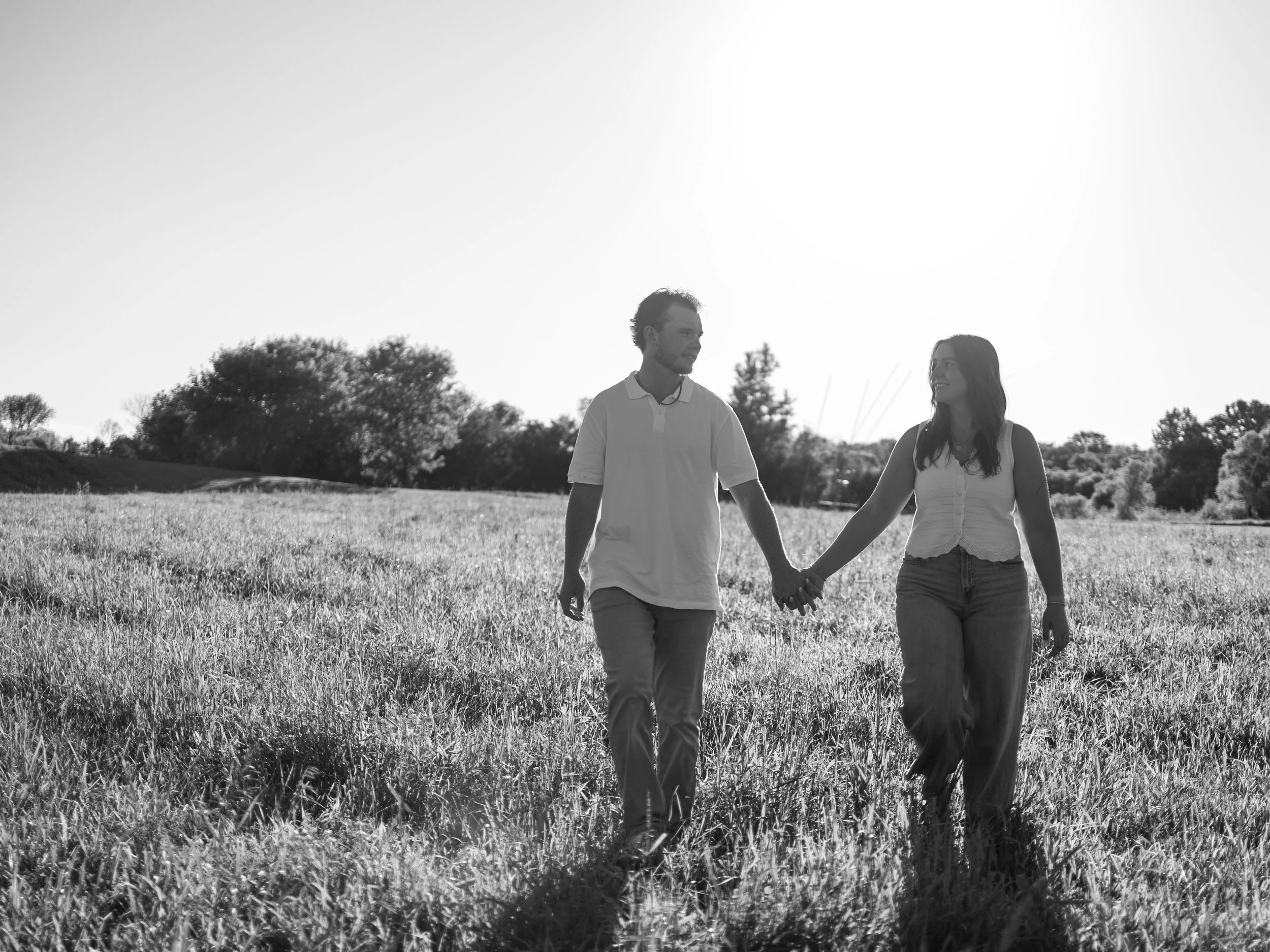 A couple holding hands and walking through a grassy field, smiling at each other, with trees and a clear sky in the background.