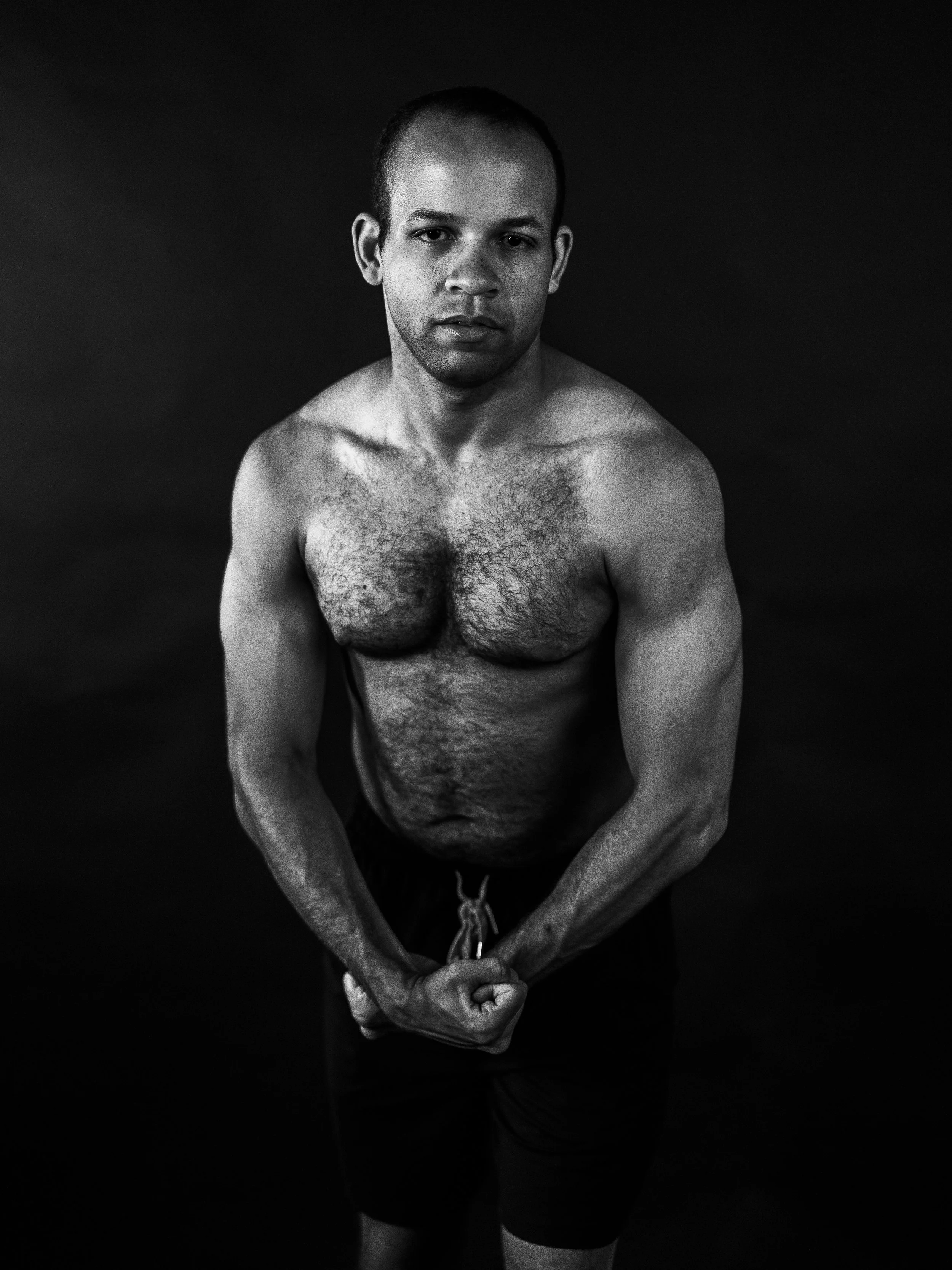 Black and white photo of a shirtless man with a serious expression, flexing his arms and clenched fists, against a dark background.