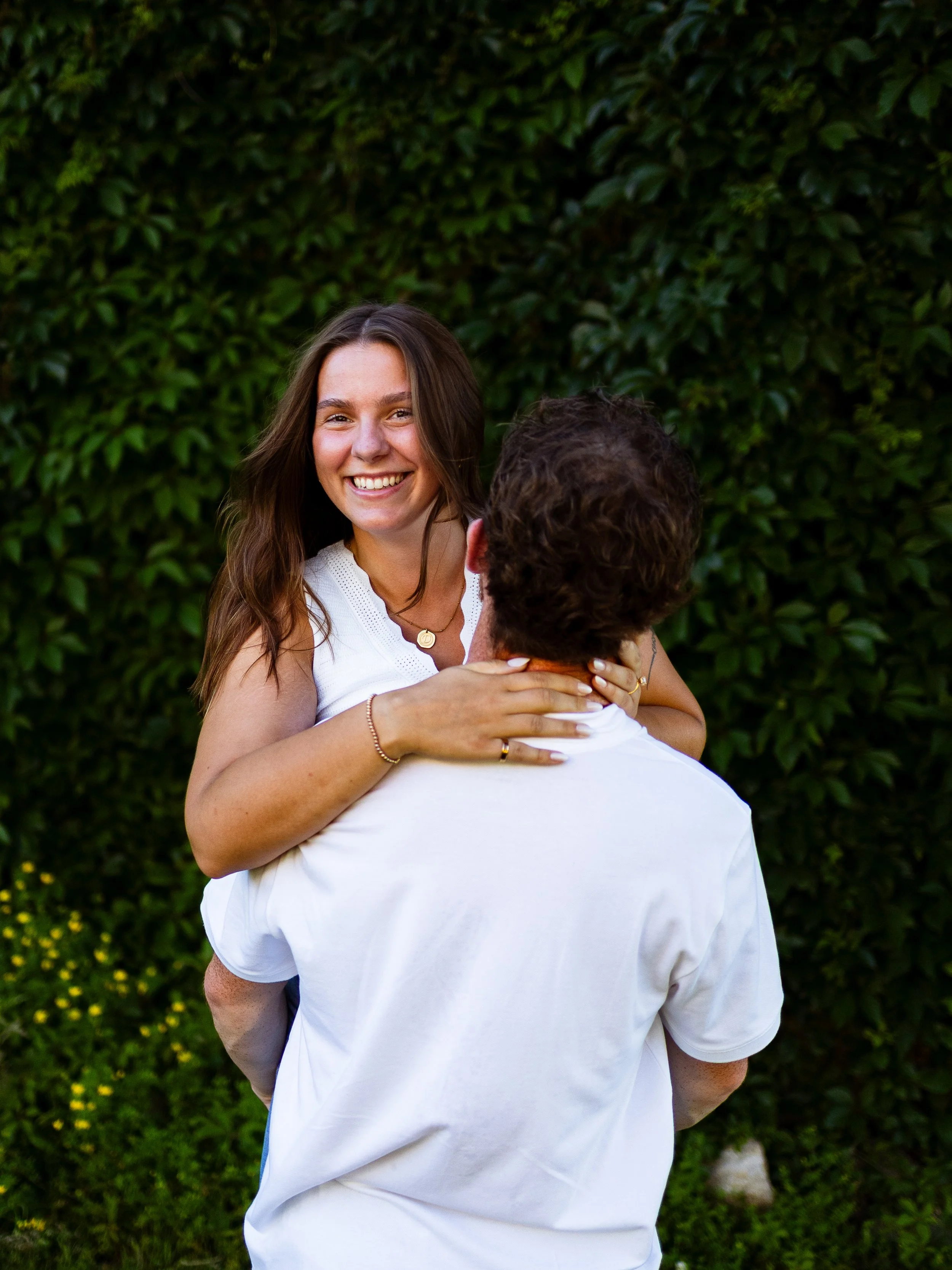 A smiling woman with long brown hair sitting on a man's shoulders outdoors, with green foliage background.