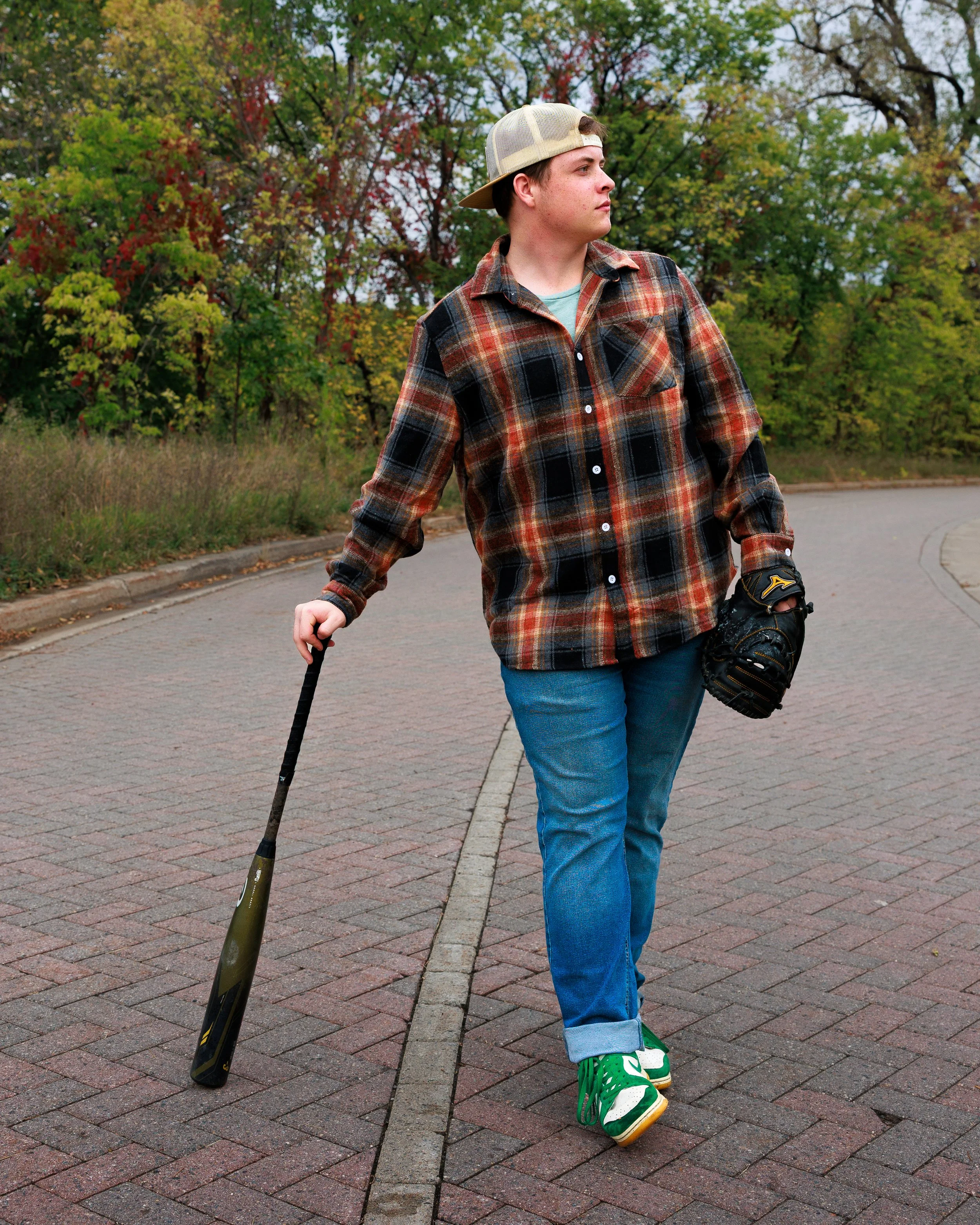 Teenager walking on a paved pathway, holding a baseball bat in his right hand, wearing a plaid shirt, jeans, green sneakers, a baseball glove on his left hand, and a backward cap, with trees in the background
