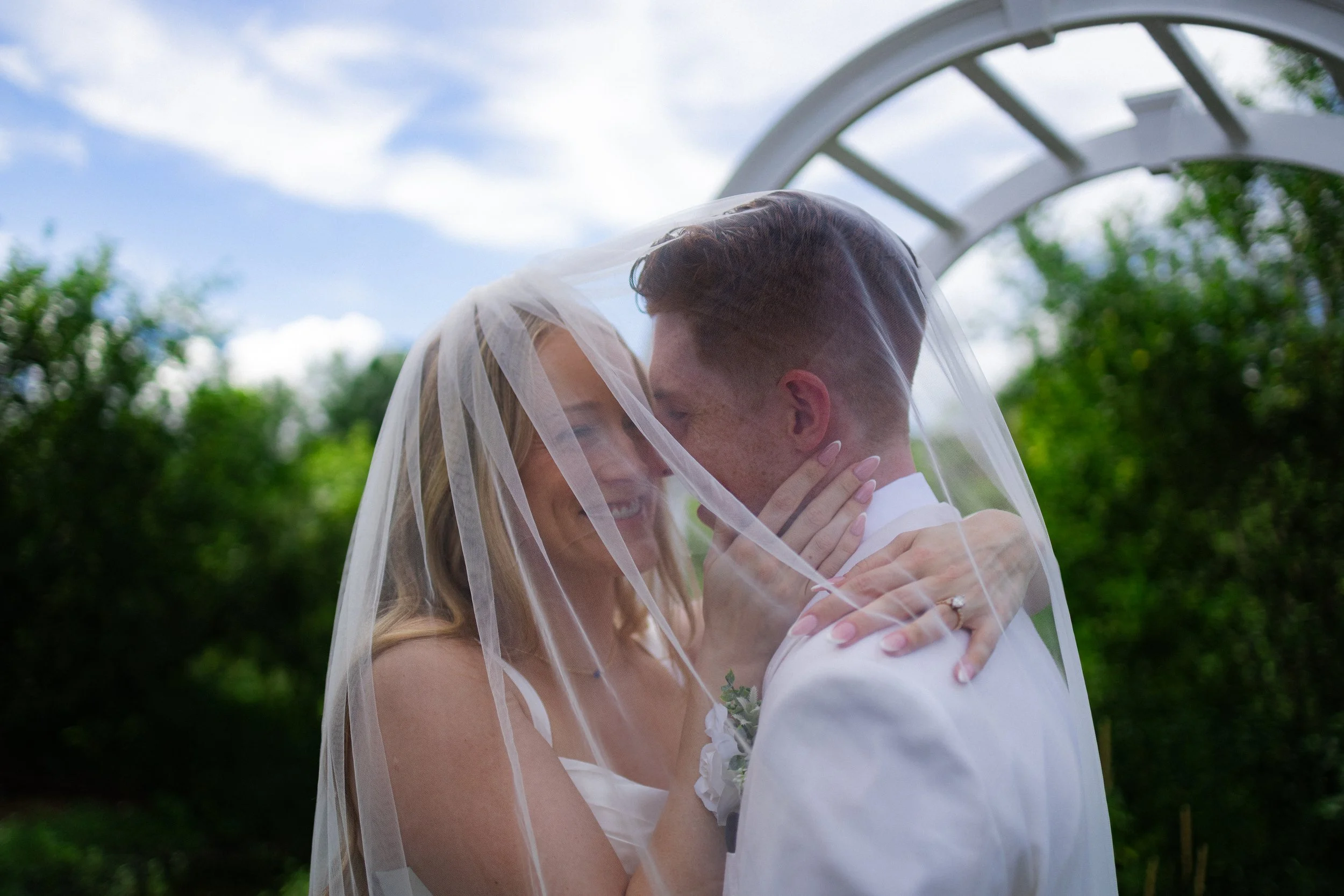 A bride and groom embracing each other outdoors under a cloudy sky with a white arch in the background, smiling and touching foreheads, with a sheer veil covering their faces.