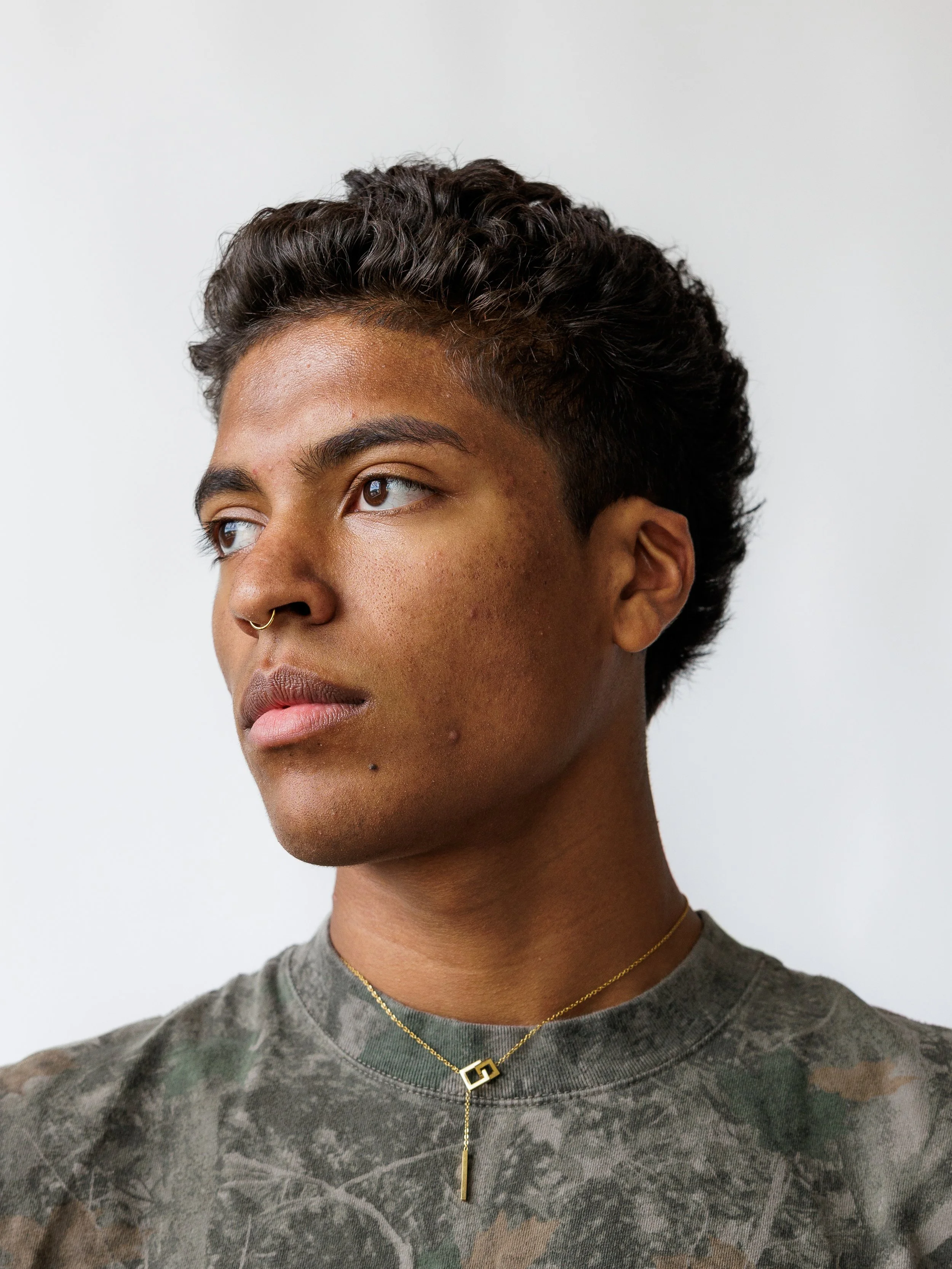 A young person with short curly dark hair, medium skin tone, and facial acne, wearing a septum piercing, a gold necklace with geometric and vertical pendant, and a camouflage-patterned shirt, looking to the side against a plain white background.