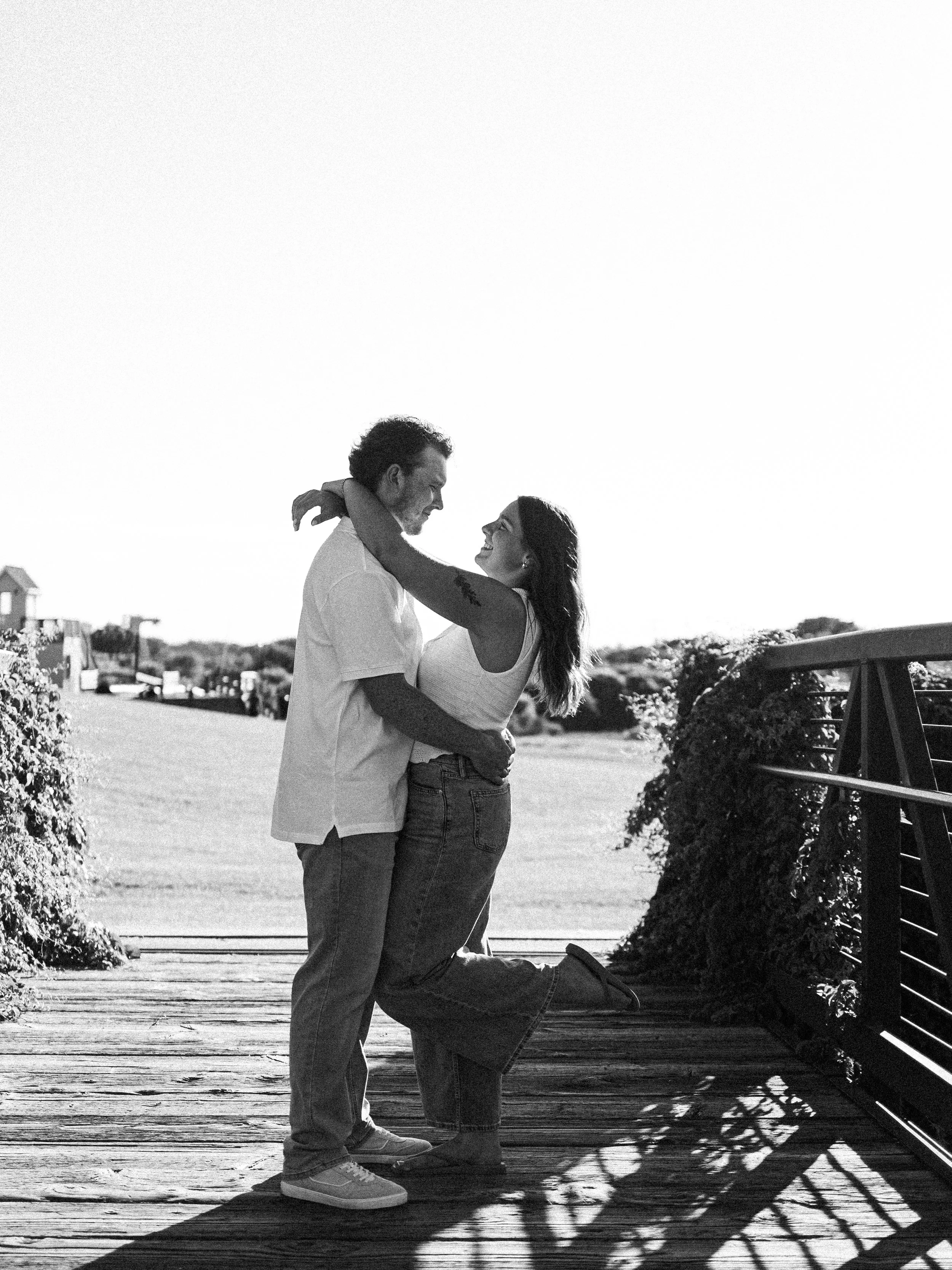 A couple embracing on a wooden bridge outdoors with a clear sky in the background.