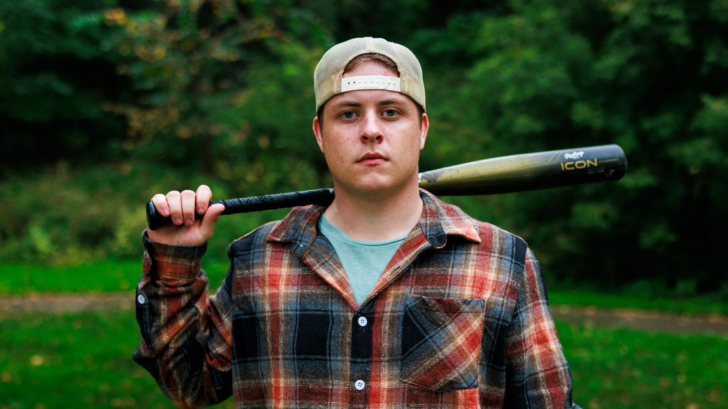 Young man holding a baseball bat over his shoulder, standing outdoors with a blurred green background, wearing a plaid shirt and a backwards baseball cap.