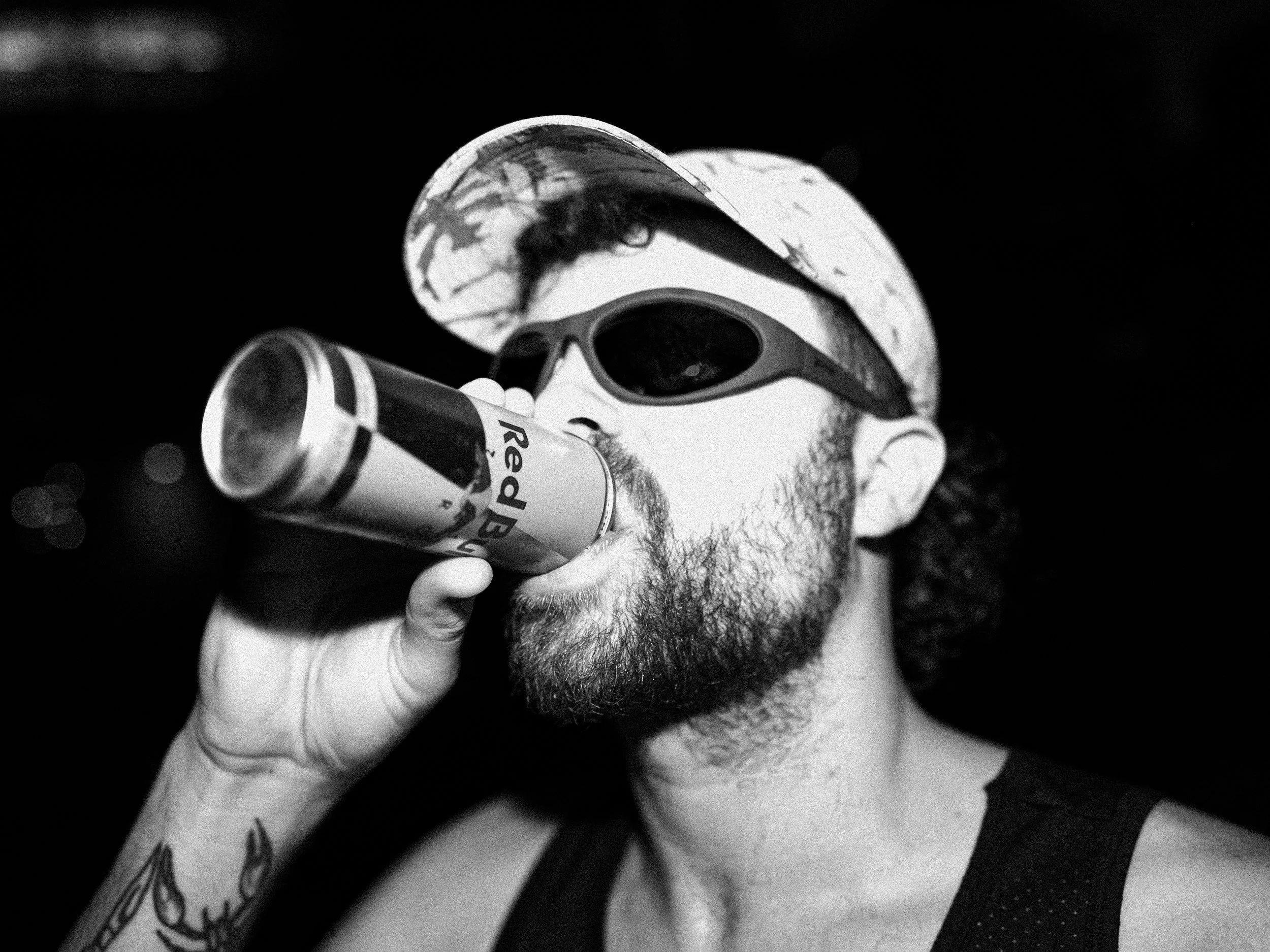 A man wearing sunglasses, a cap, and a tank top is drinking from a can of Red Bull energy drink.