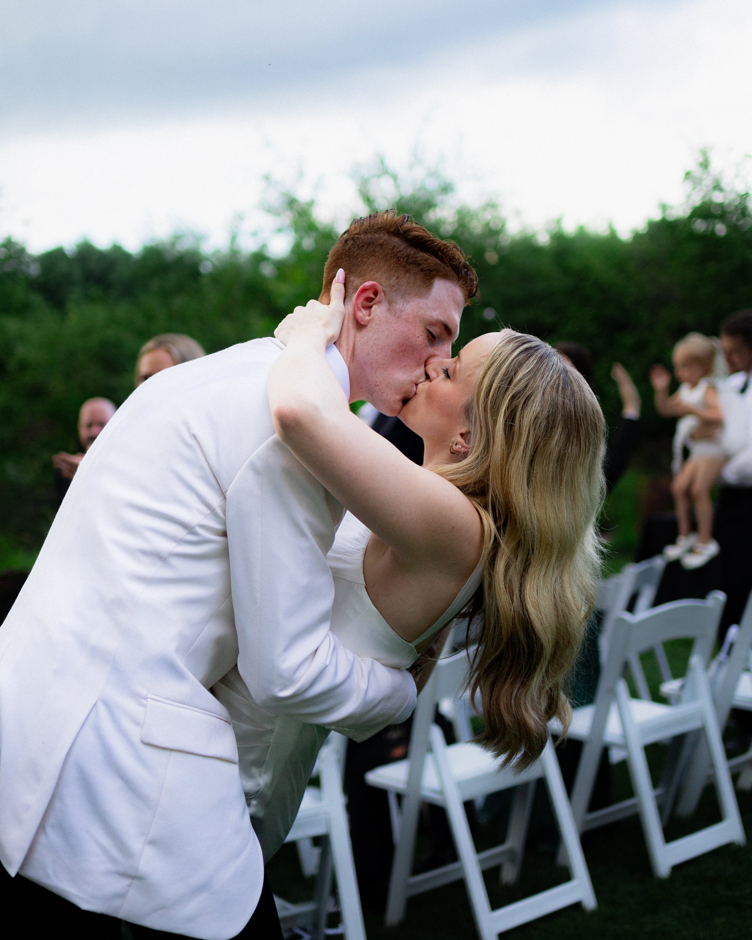Couple sharing a kiss at an outdoor wedding ceremony, surrounded by guests and white chairs, with a cloudy sky and greenery in the background.