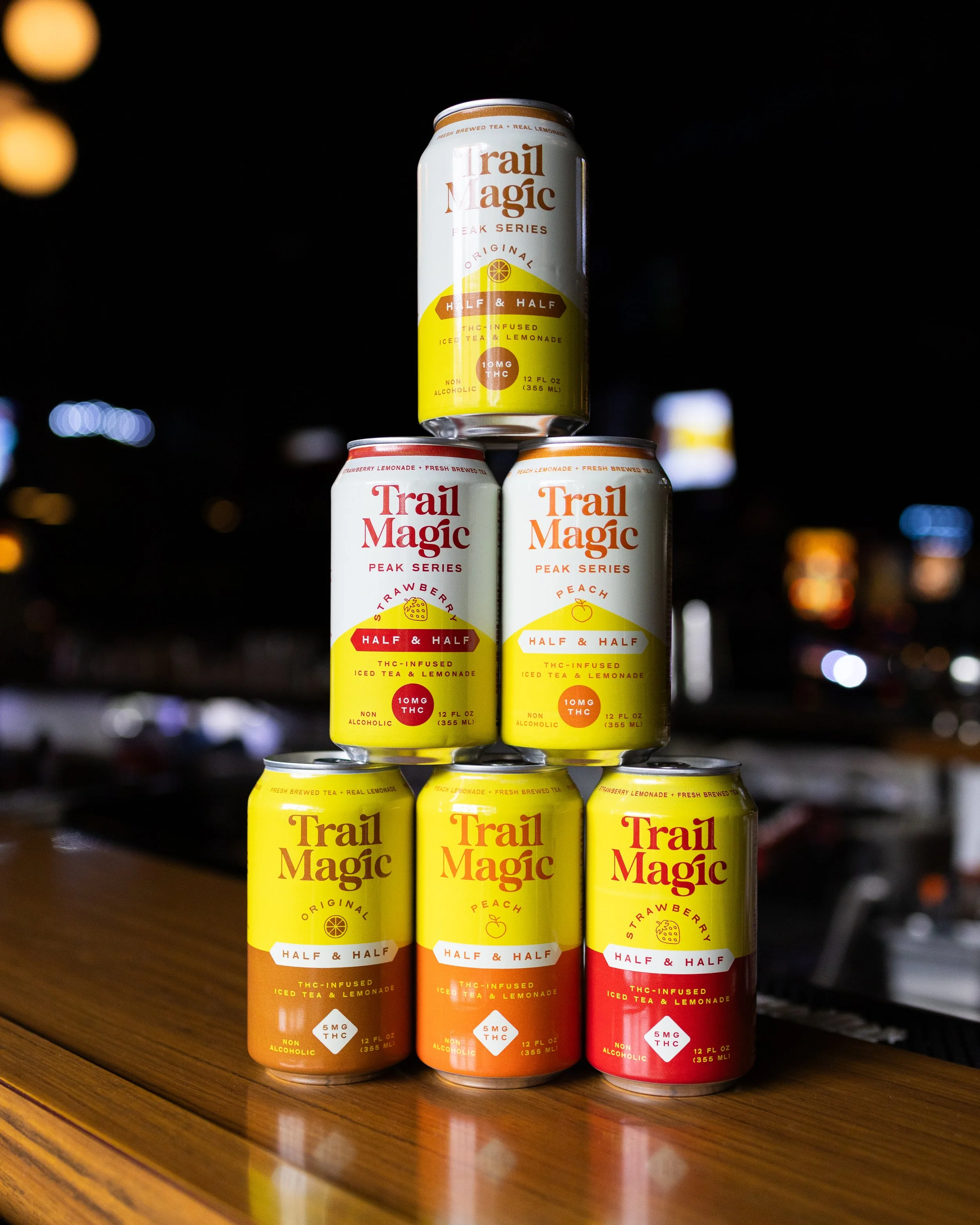 Six cans of Trail Magic THC-infused iced tea and lemonade drinks arranged in a pyramid on a wooden surface at night, with a dark background and blurred lights.