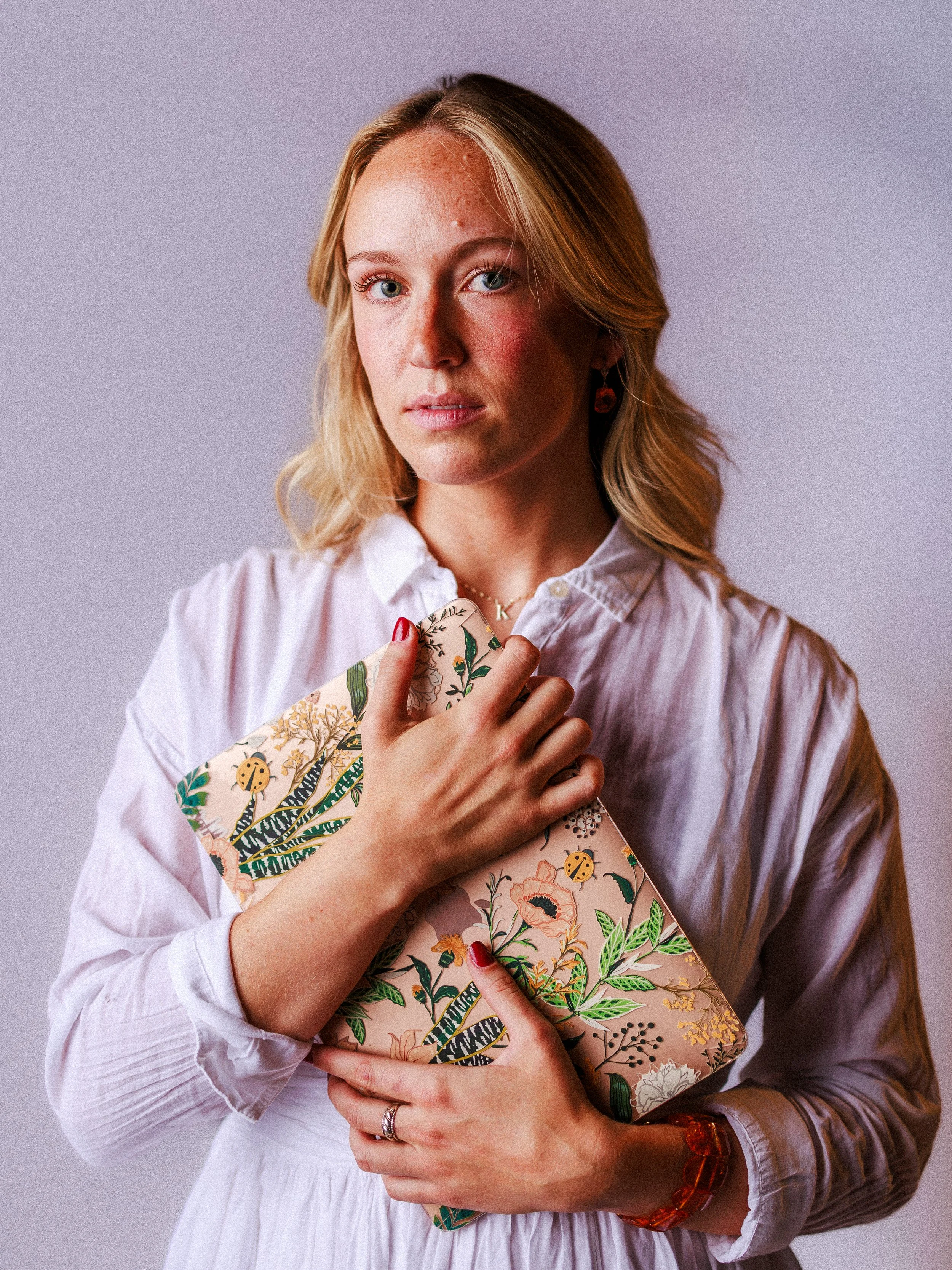 A young blonde woman wearing a white blouse holding a floral patterned notebook close to her chest.