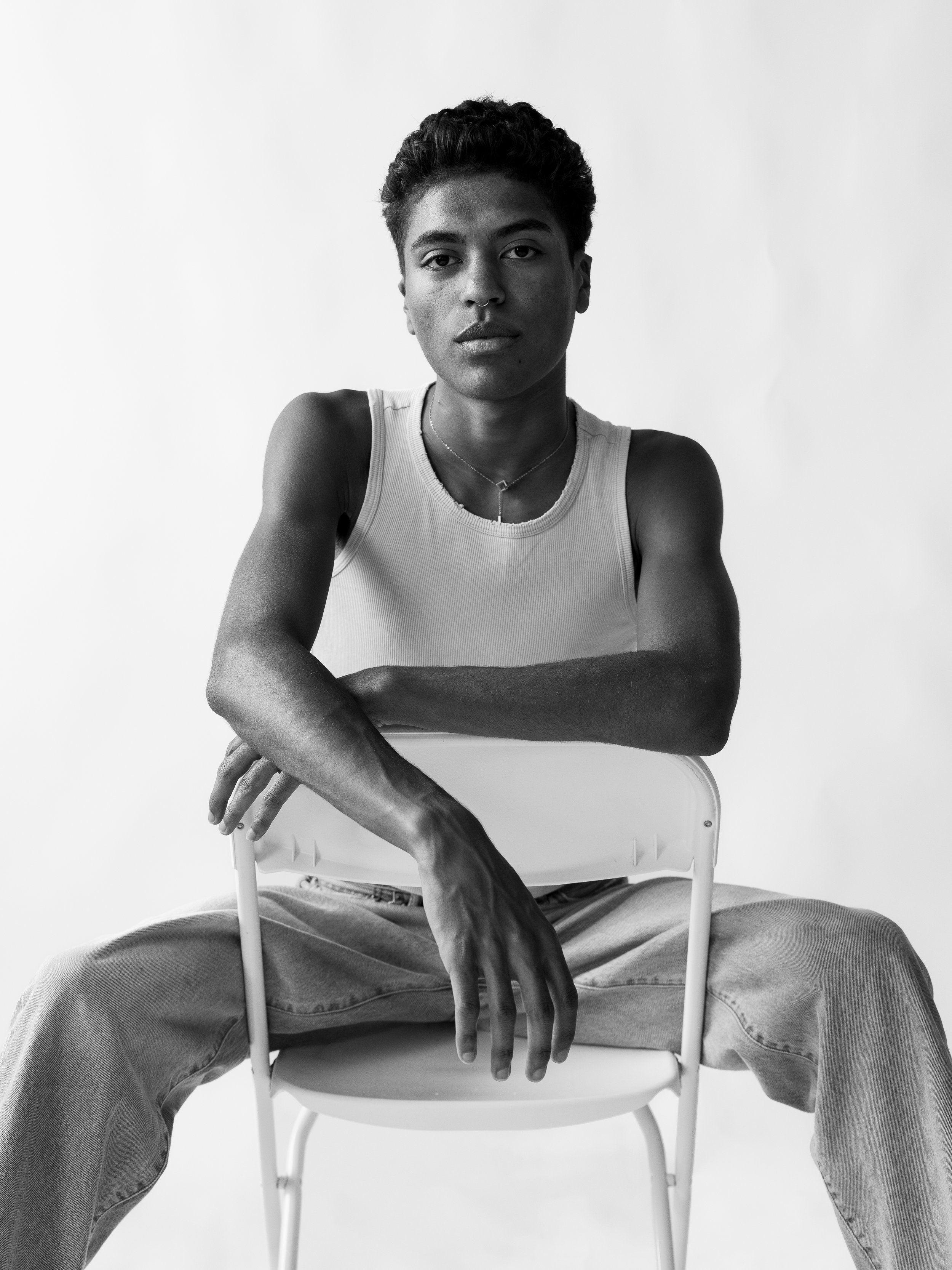 Black and white photo of a young person sitting on a chair with legs wide apart, wearing a sleeveless shirt and jeans, looking directly at the camera with a serious expression.