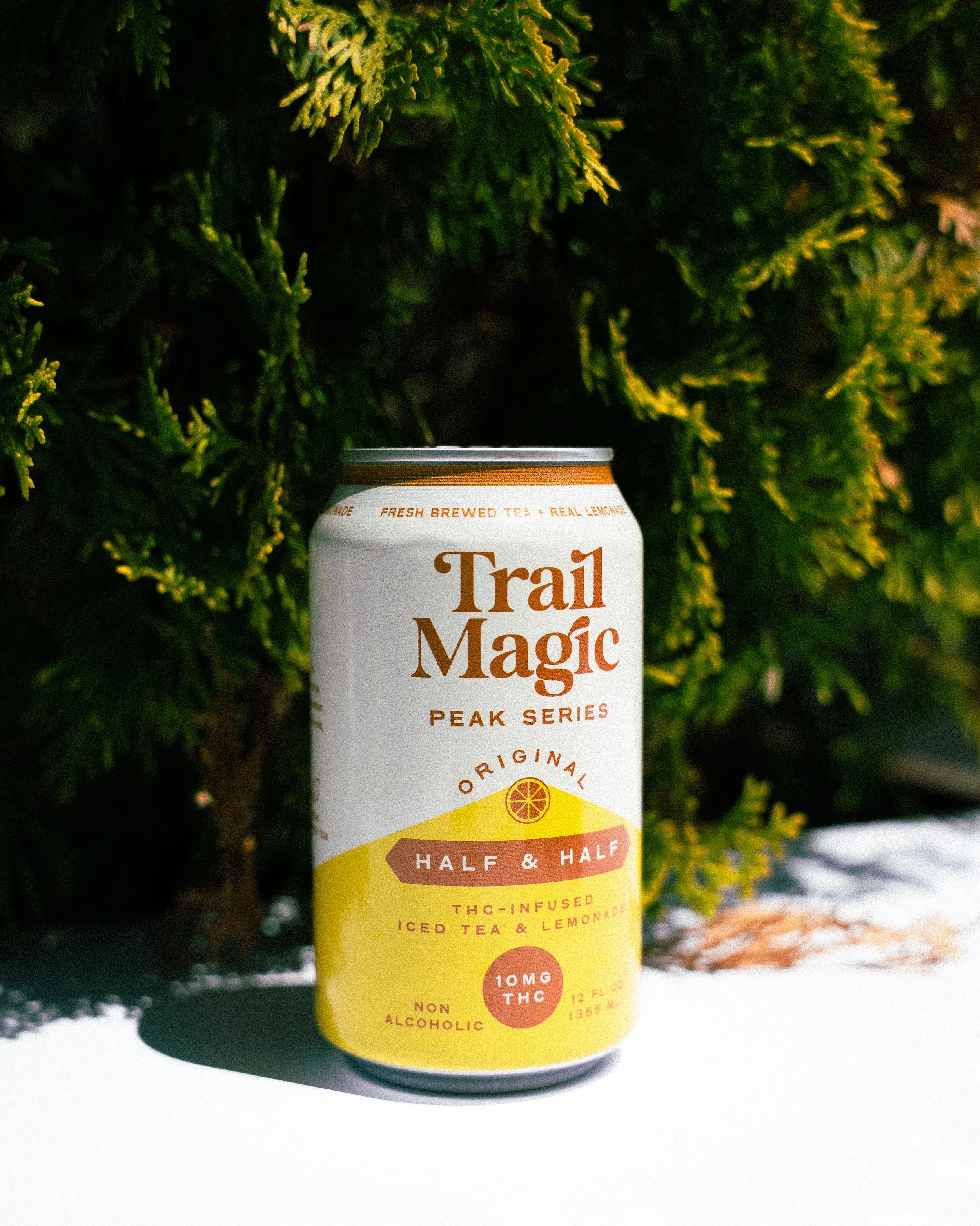 A can of Trail Magic Peak Series iced tea with lemon flavor, placed outdoors on a white surface against green pine tree branches.