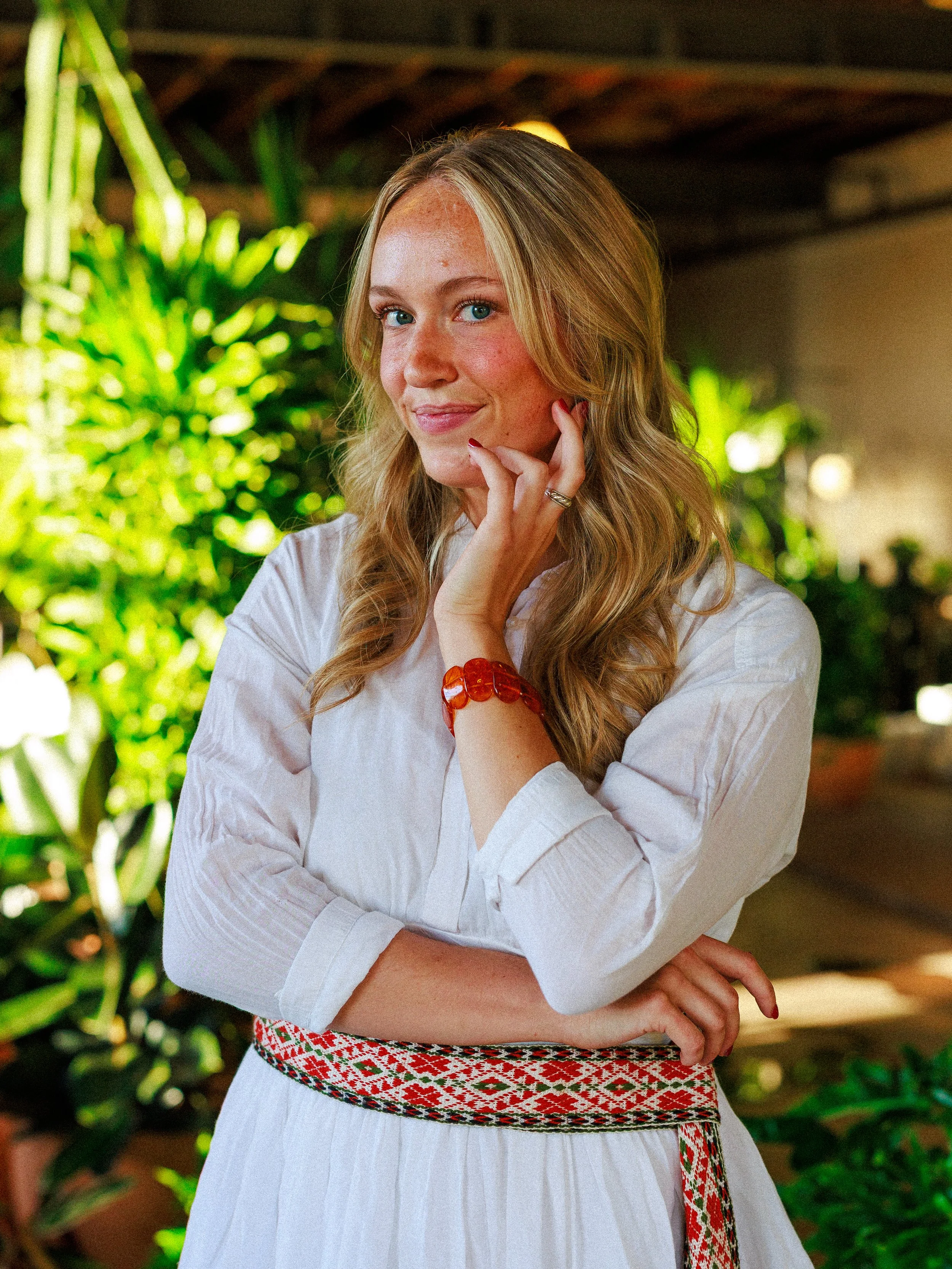 A woman with long blonde hair, wearing a white blouse, red bracelet, and a colorful embroidered belt, standing in front of green plants, smiling softly with her hand touching her cheek.