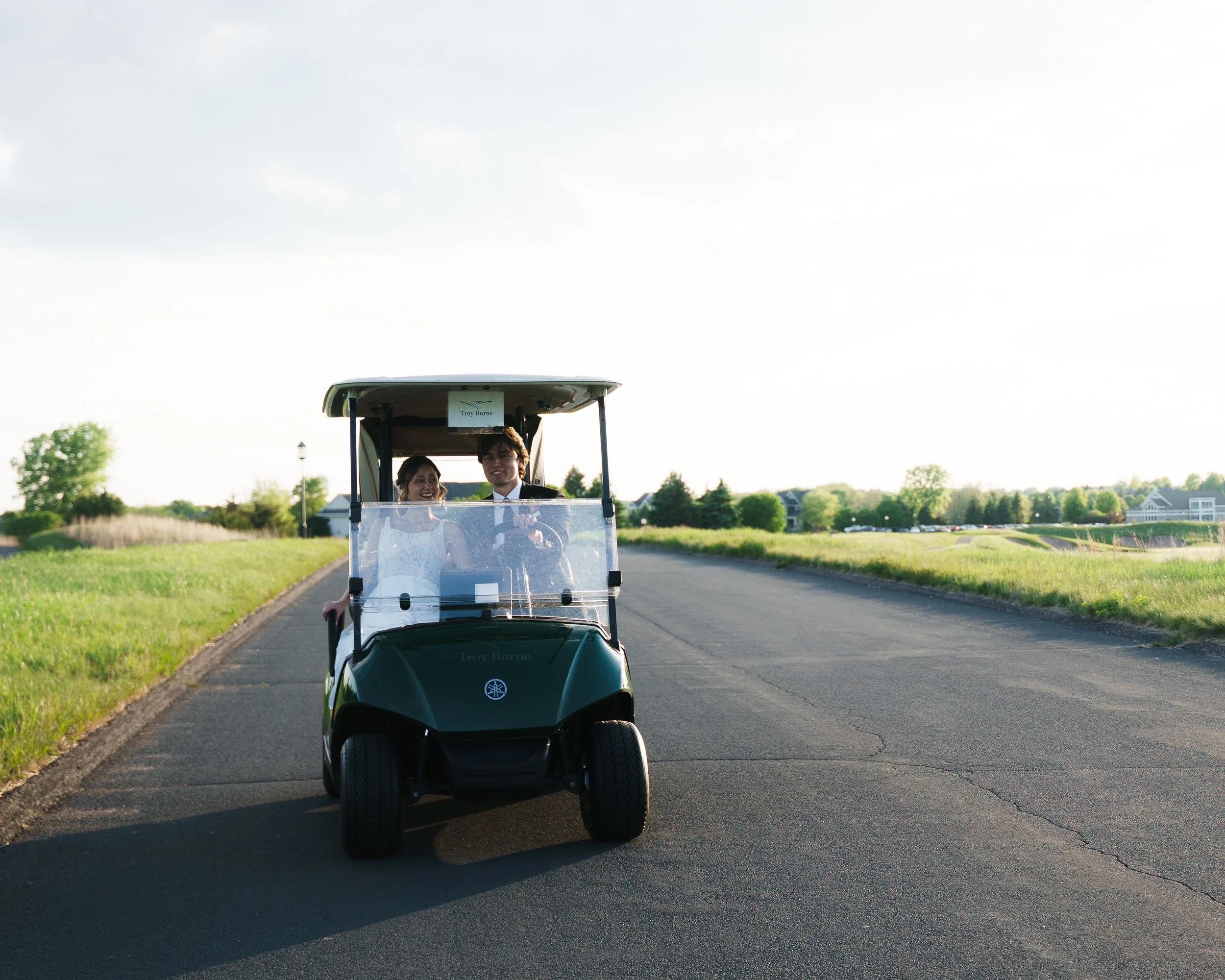 A couple riding in a golf cart on a paved path through a green park in the late afternoon.