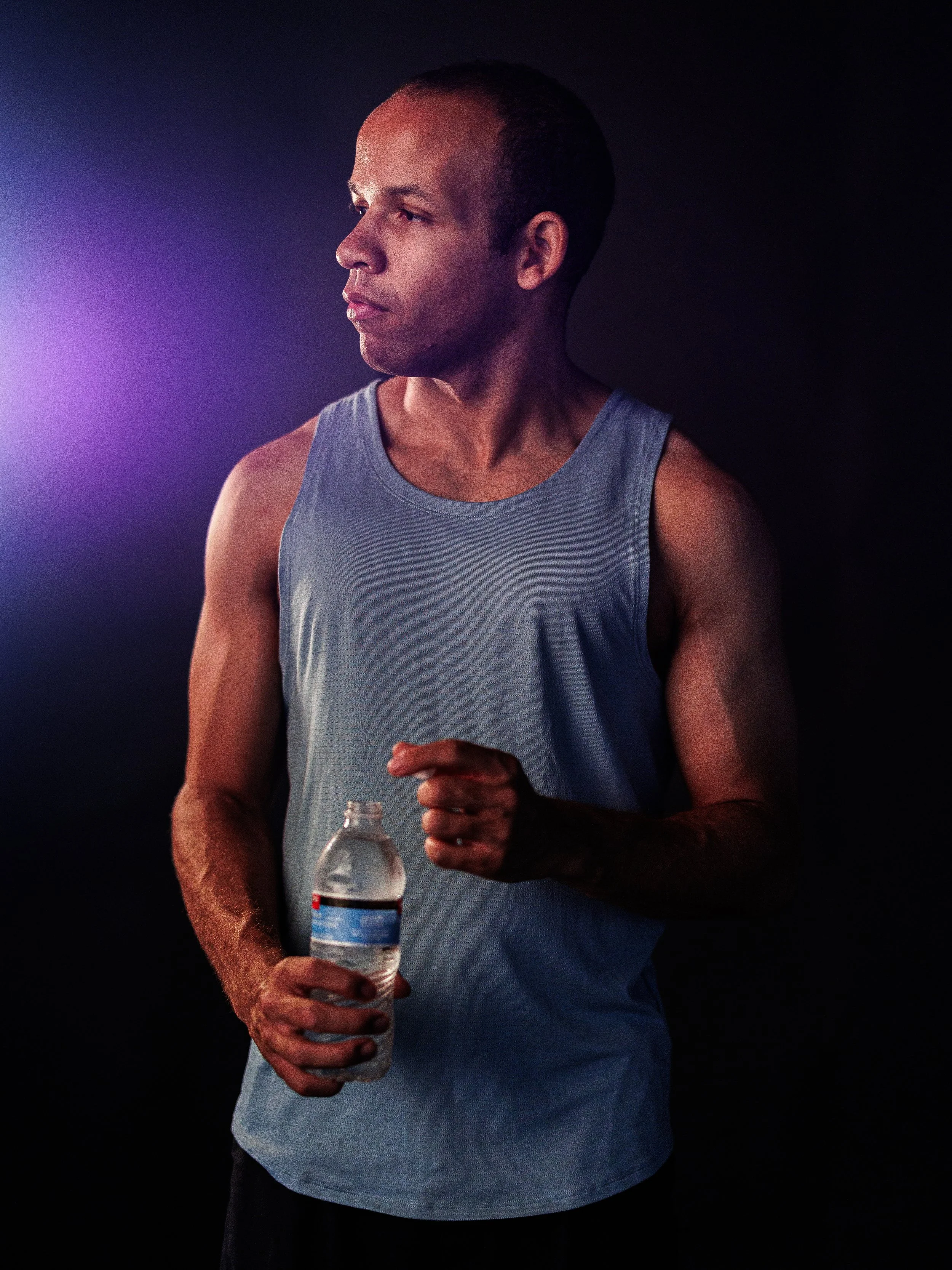 Man in sleeveless gray shirt holding a water bottle, looking to the side against a dark background with purple light on the left.