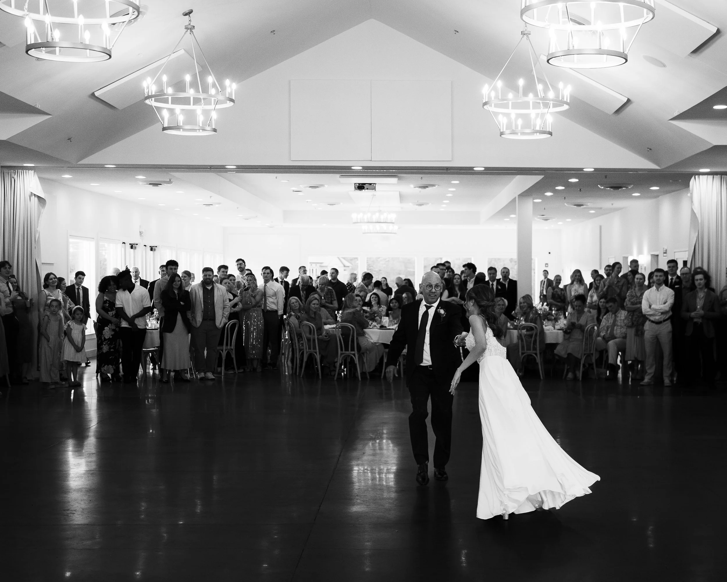 Black and white photo of a wedding reception with a bride dancing with an older man on the dance floor, surrounded by guests seated at tables and standing around the perimeter in a brightly lit hall with chandeliers.